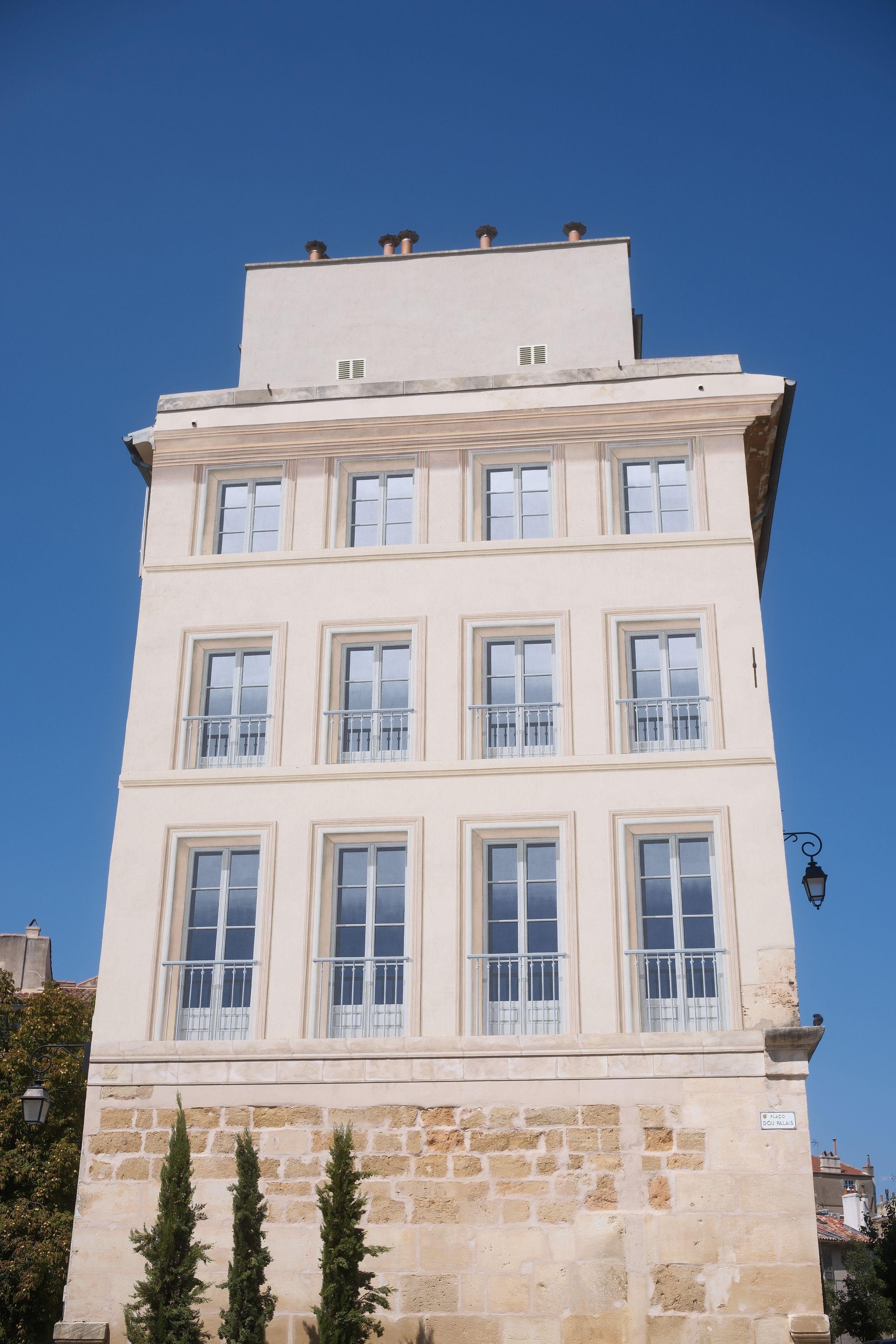 Photo format portrait montrant la façade d’un immeuble avec 4 fenêtres par étage, sous un ciel bleu. Les fenêtres sont des trompe-l’œil très réussis