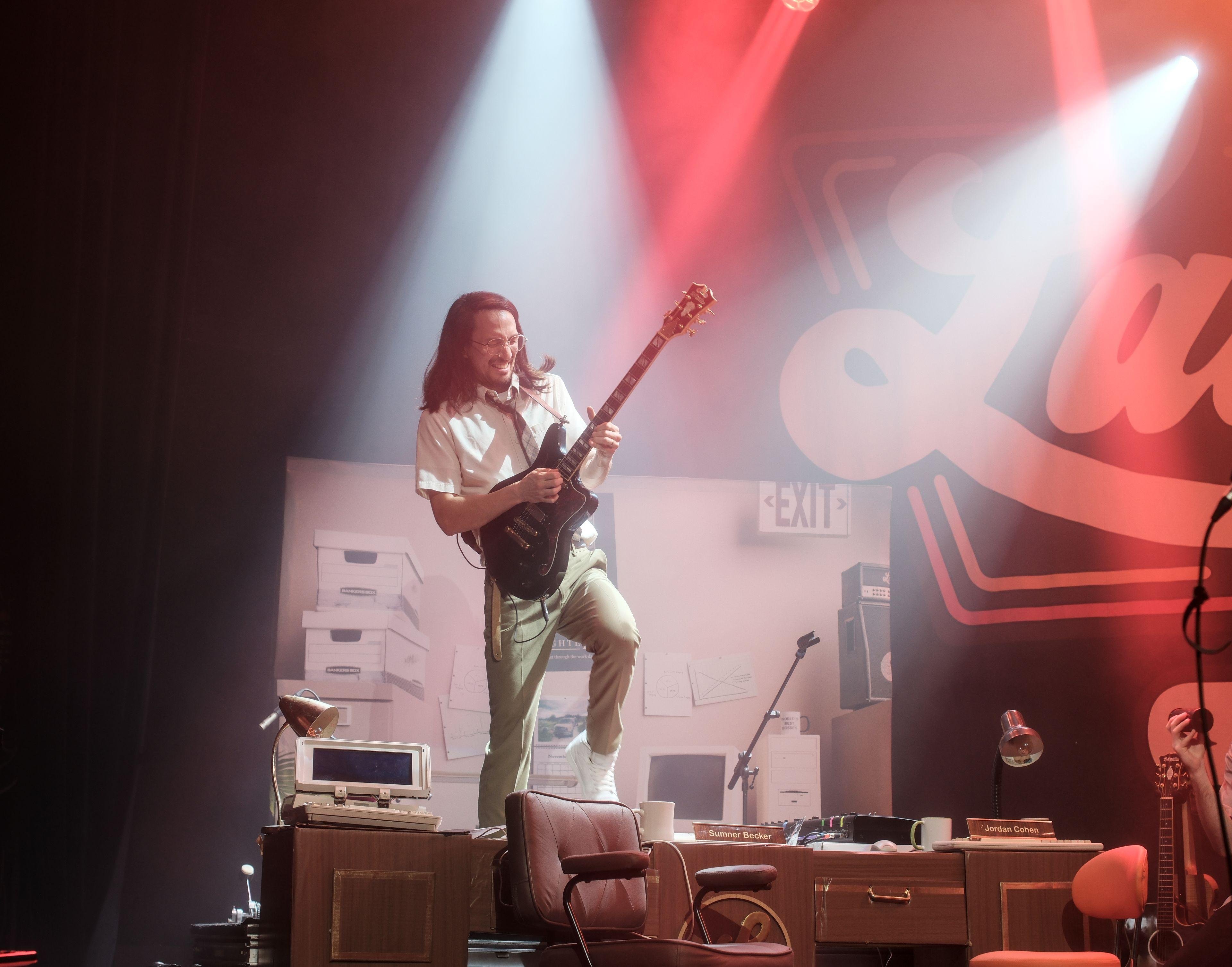Photo format portrait d’un concert où on voit un homme aux cheveux longs et noir debout sur une table de bureau en train de faire un solo de guitare, dans une pose dynamique, éclairé par deux spots blancs sur une scène éclairée en rouge/orange