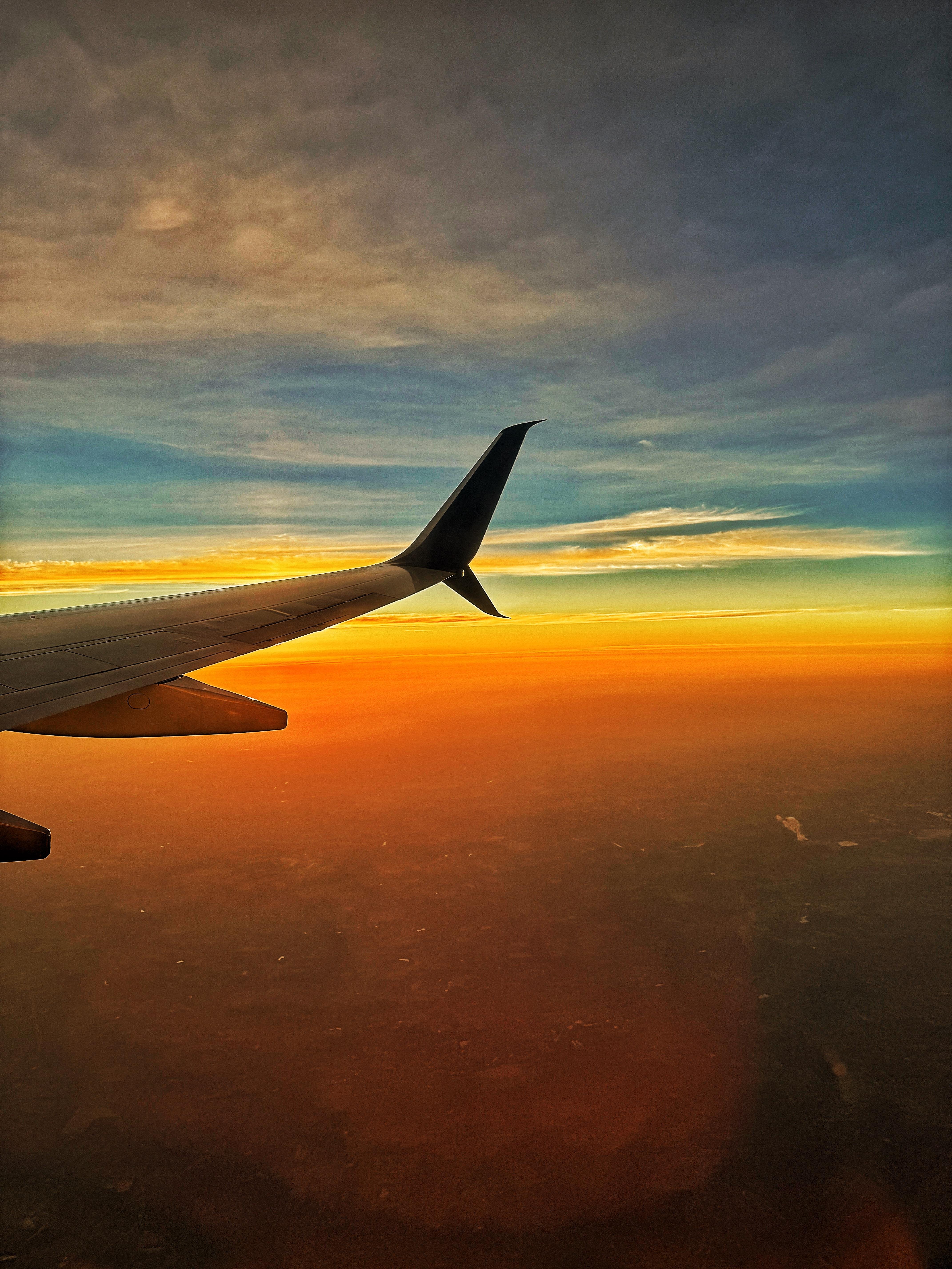 The image captures a view from an airplane window during sunset. The airplane's wing is prominently visible in the foreground, extending from the bottom left corner towards the center of the image. The wing is silhouetted against a vibrant sky, which transitions from a deep blue at the top to warm hues of orange and yellow near the horizon. The clouds in the sky are scattered, with some areas appearing darker, possibly due to the setting sun. Below the airplane, the landscape is bathed in a reddish-orange glow, suggesting the sun is just below the horizon. The overall scene conveys a sense of tranquility and the beauty of nature from an aerial perspective.