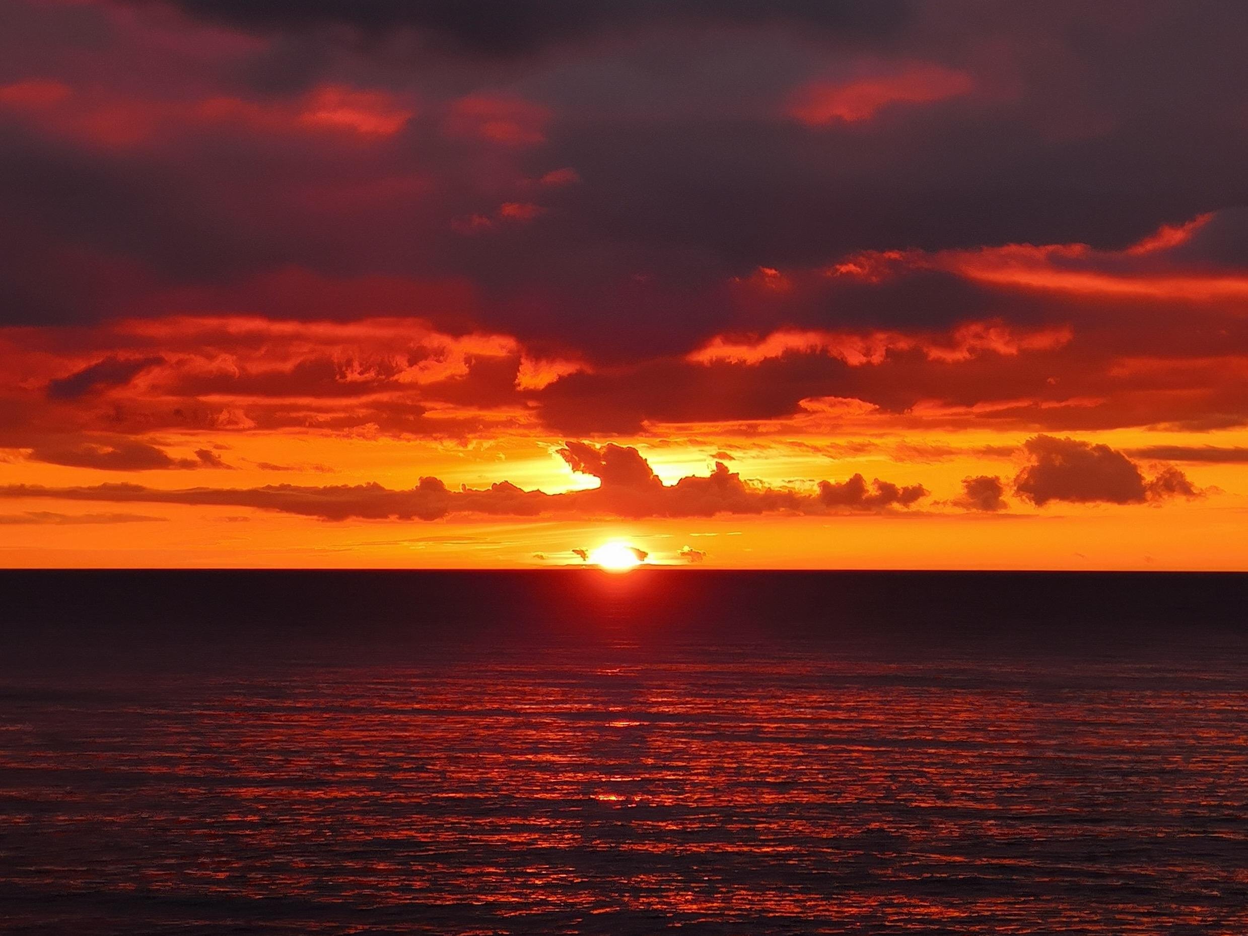 Sun setting at the horizon over a calm ocean, with dark layered clouds above and orange-red light reflecting across the water.