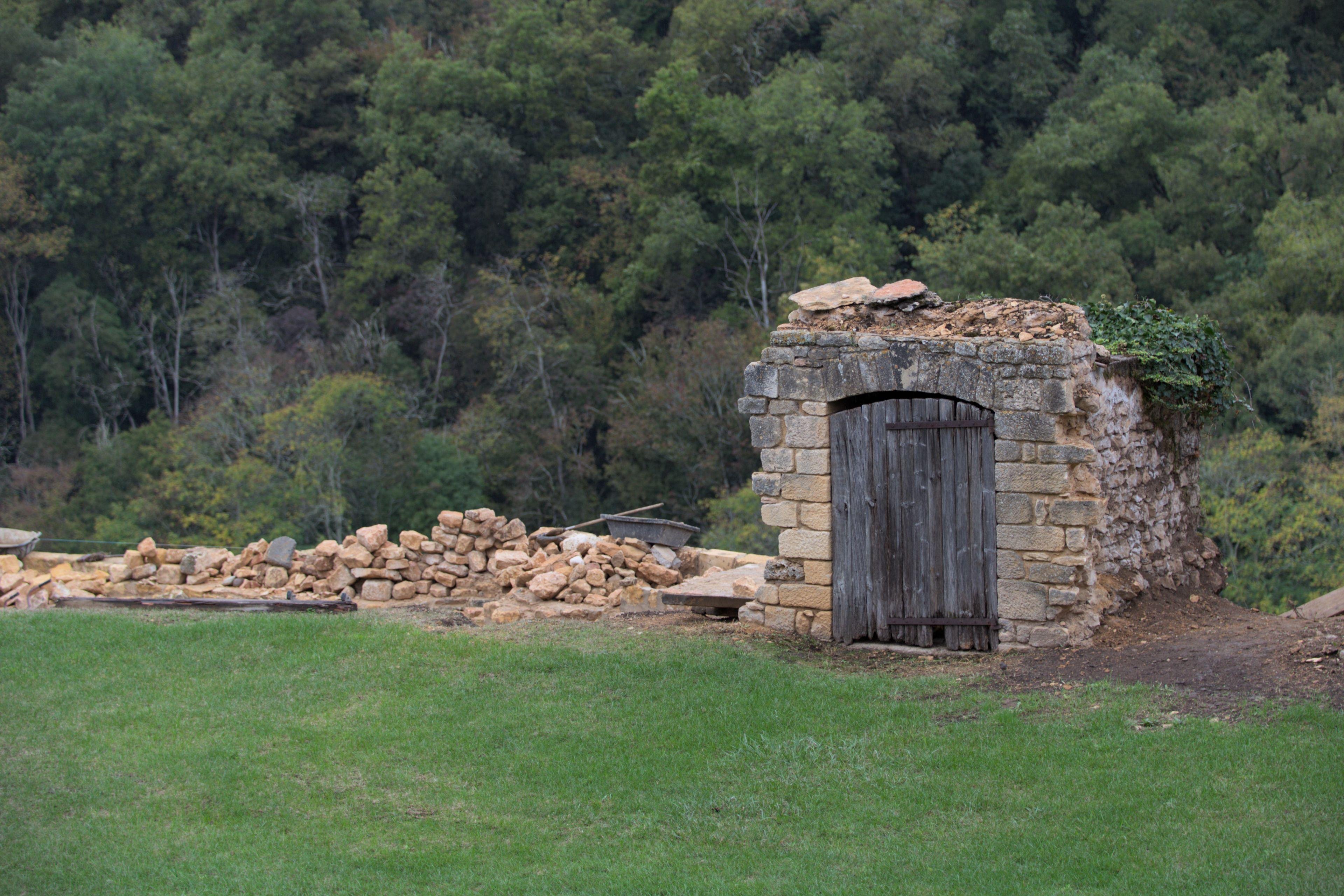Old small stone building with a wooden door which was part of something bigger, surrounded by scattered stones and green grass, with a forest in the background.