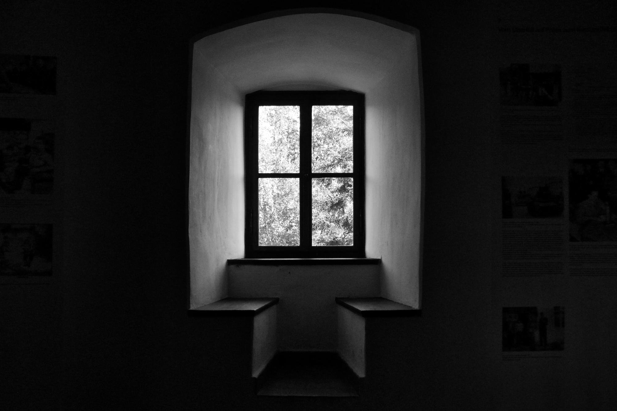 Recessed arched window with four panes centered in a dark room, bright light and tree foliage visible outside; built-in bench ledges on both sides of the window alcove.