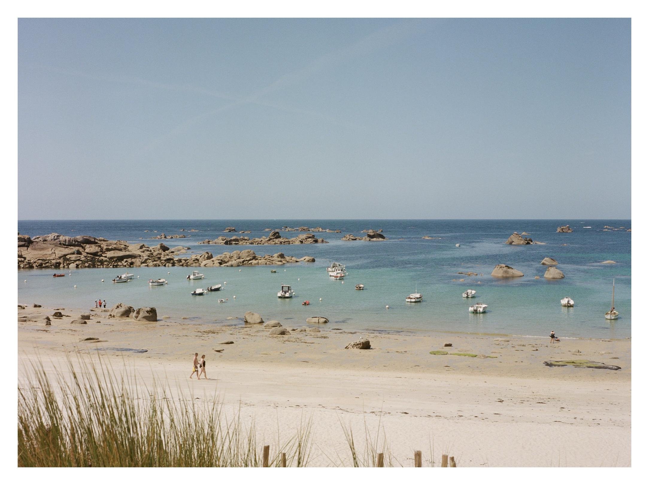 Wide sandy beach with dune grass in the foreground, two people walking near the shoreline, and a calm turquoise bay dotted with small anchored boats and rocky outcrops under a clear sky.