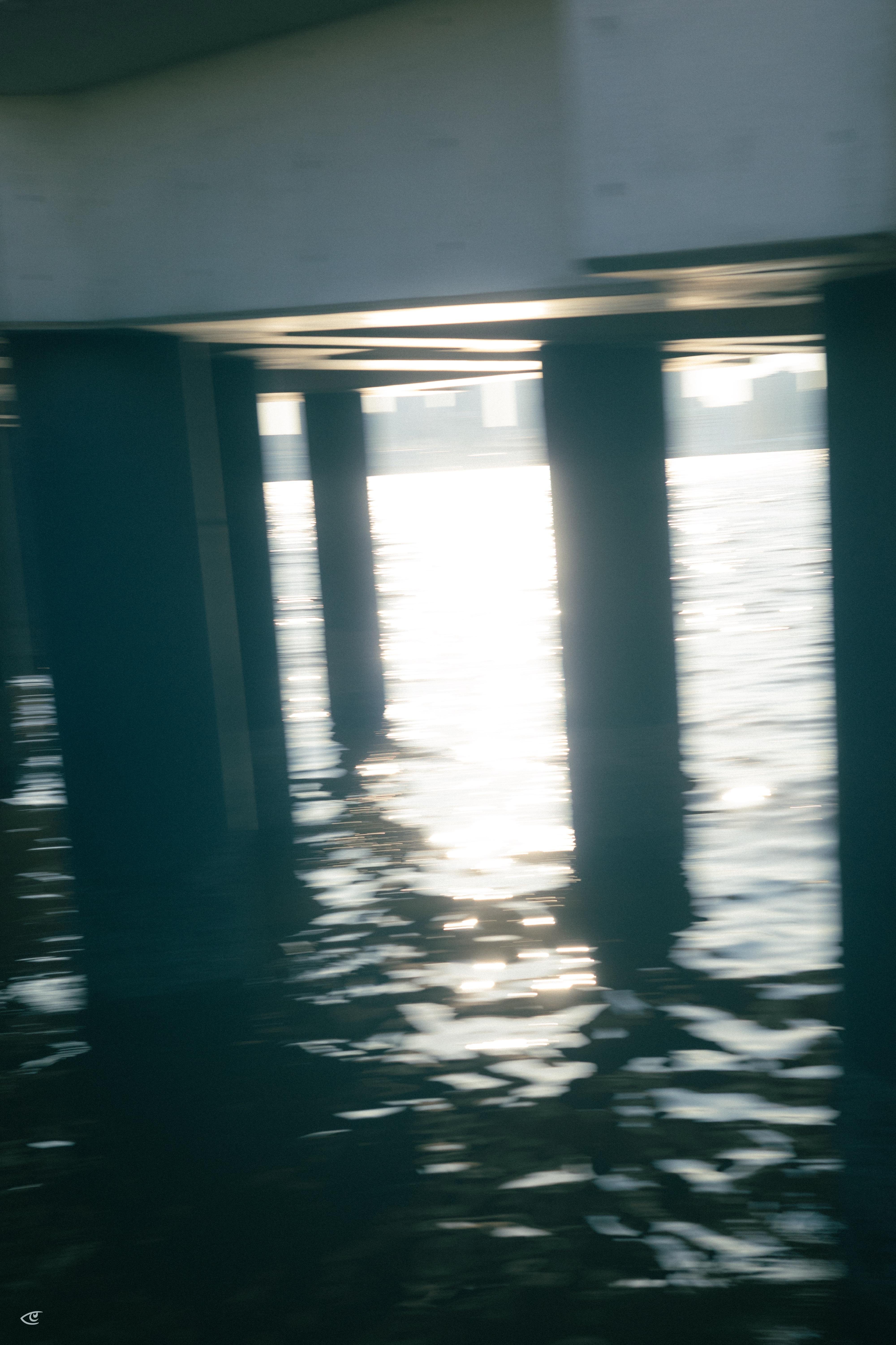 View from beneath a pier with dark vertical support pilings over rippling water, bright sunlight reflecting in a vertical strip near the center, and a faint city skyline in the distance.