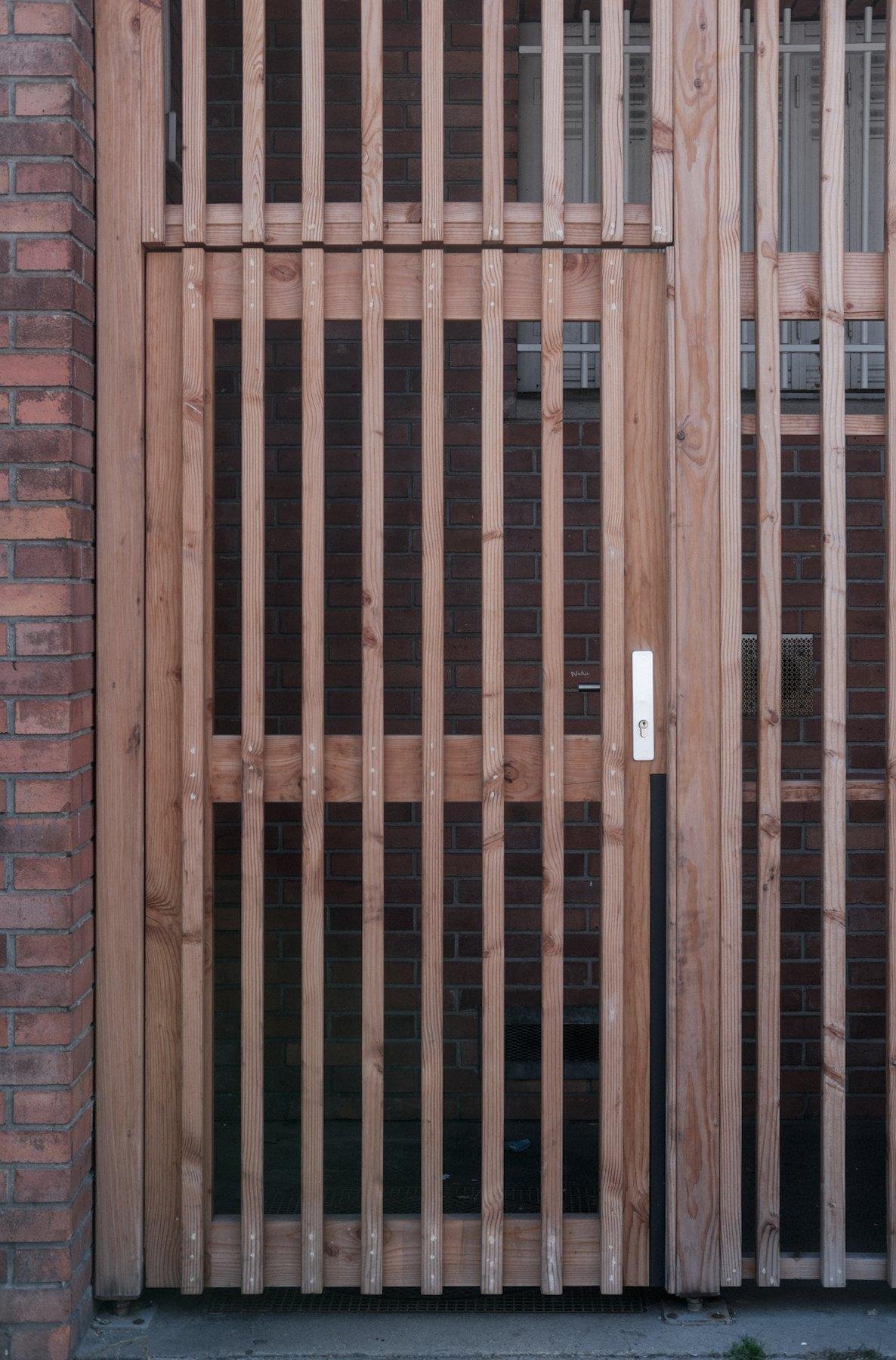 Photo verticale. Une porte en grille de bois aux barreaux verticaux qui donne sur un passage aux murs en brique