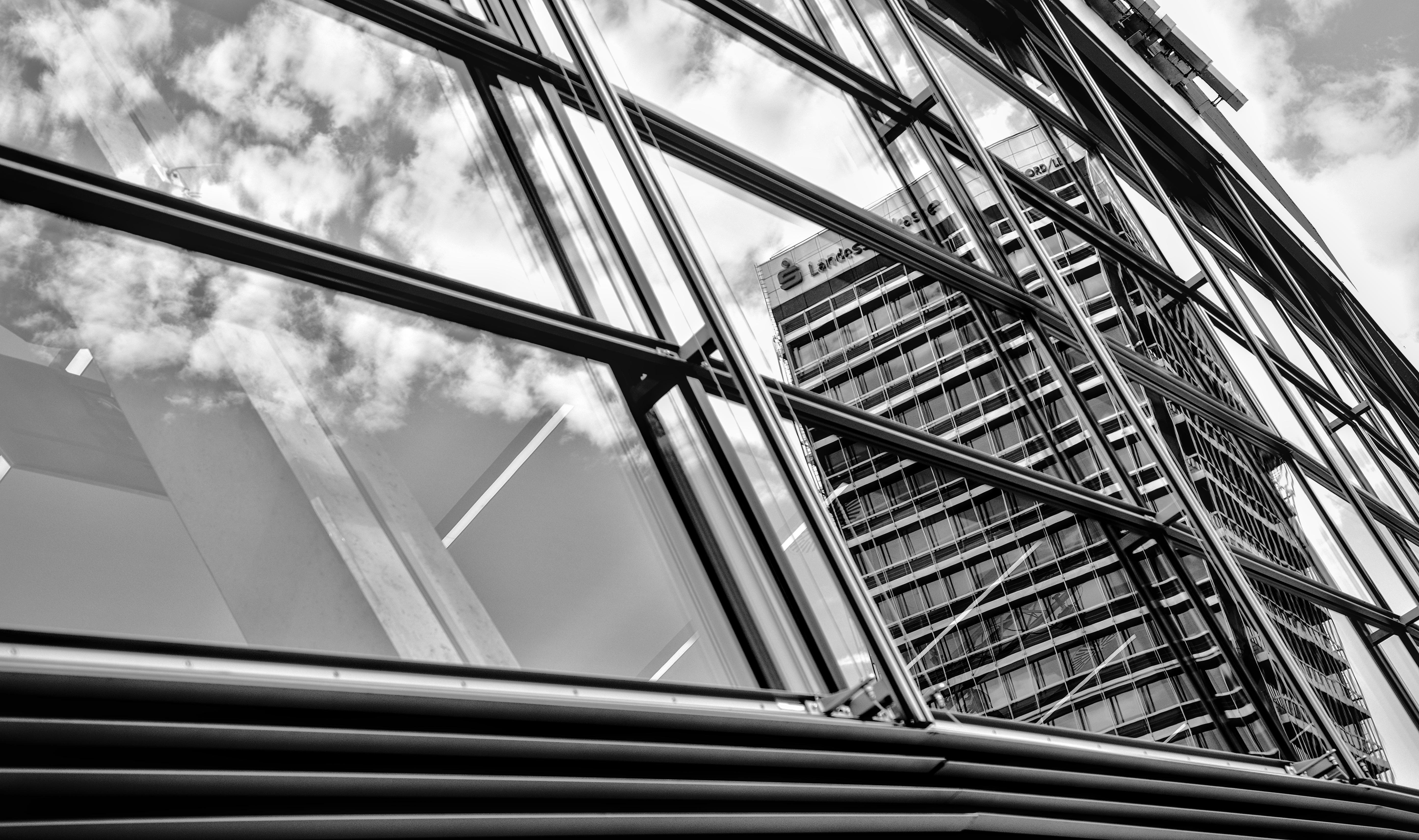 Low-angle black-and-white view of a modern glass-and-steel building facade with diagonal beams and window reflections of clouds, with a tall office tower visible in the background.