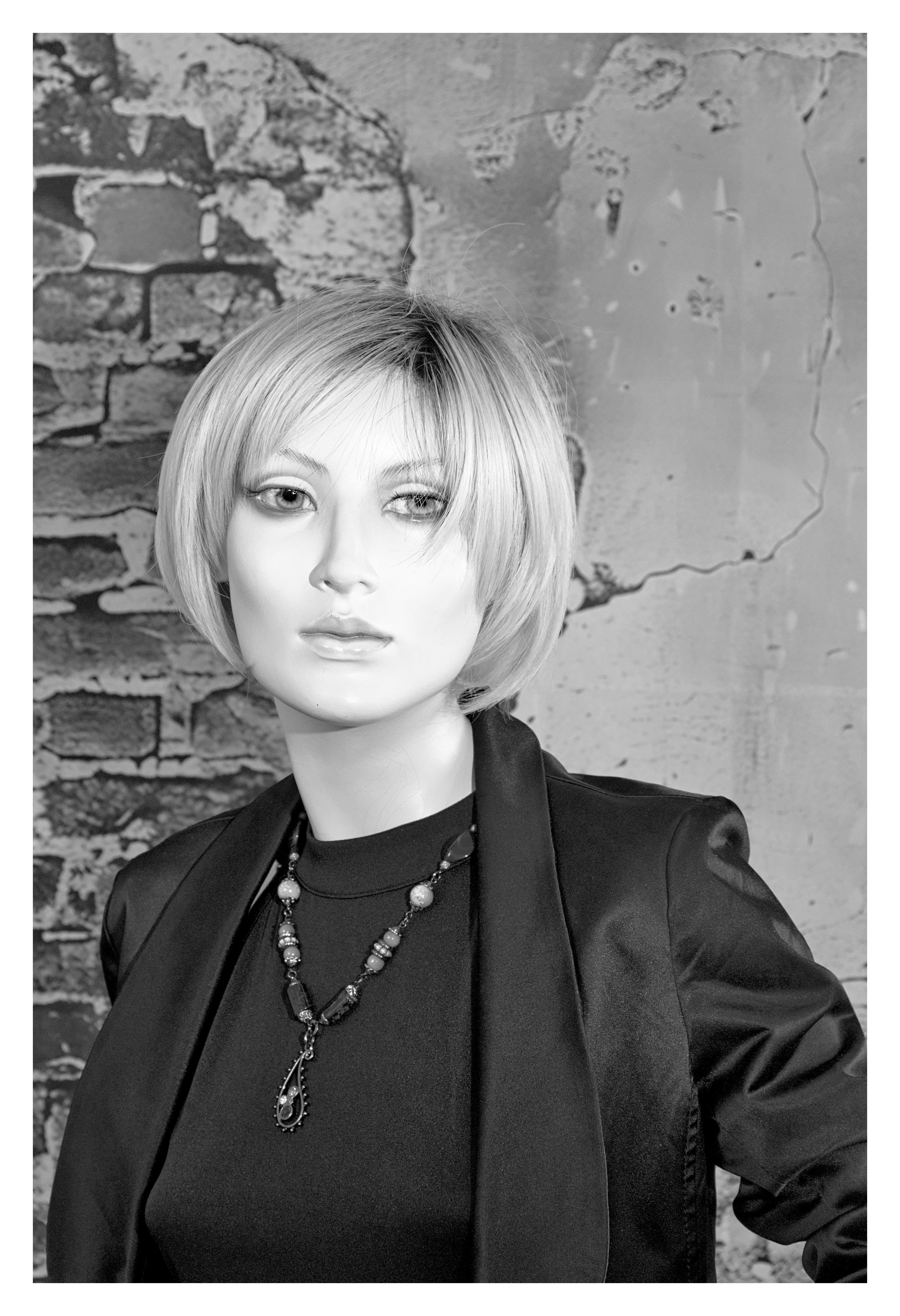 The image shows a mannequin with short, styled hair, wearing a dark outfit with a jacket. The mannequin is also wearing a decorative necklace. The background includes a textured, cracked wall with a brick pattern. The photo is in black and white.
