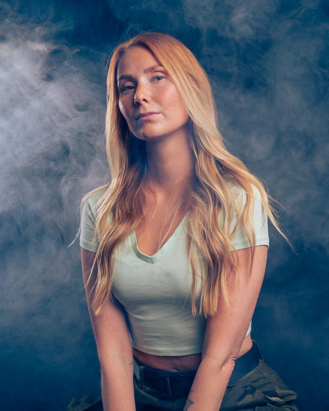 The image shows a person with long blonde hair wearing a light-colored shirt and dark pants. The background has a misty or smoky effect, creating a dramatic atmosphere.