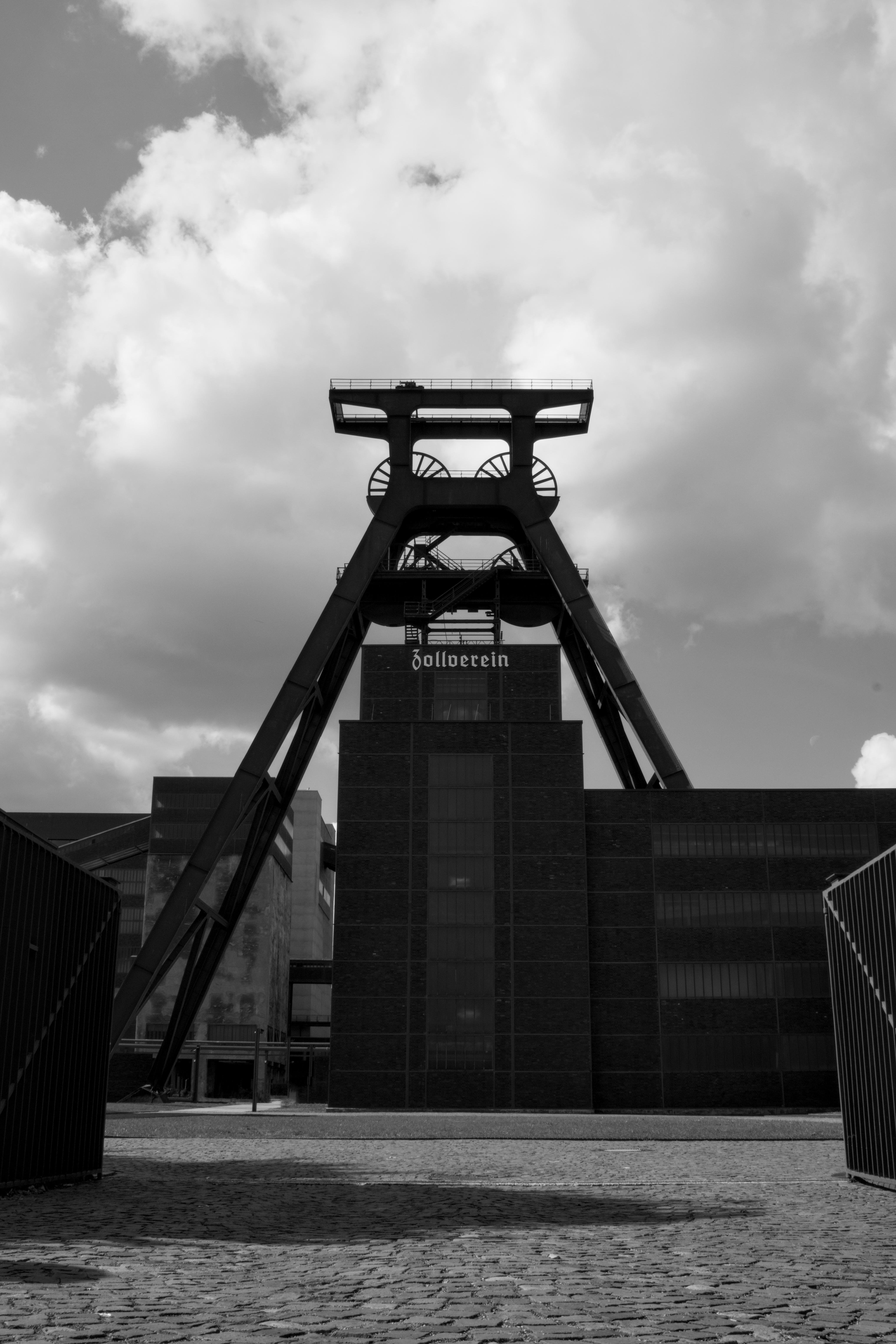 Tall industrial structure with prominent angular beams and wheels, labeled "Zollverein" at the top. Surrounding buildings have a modern, geometric design. Sky is filled with fluffy clouds, adding a dynamic backdrop. Cobbled ground in the foreground enhances the historical industrial setting. The image is in black and white, emphasizing contrasts and architectural details.