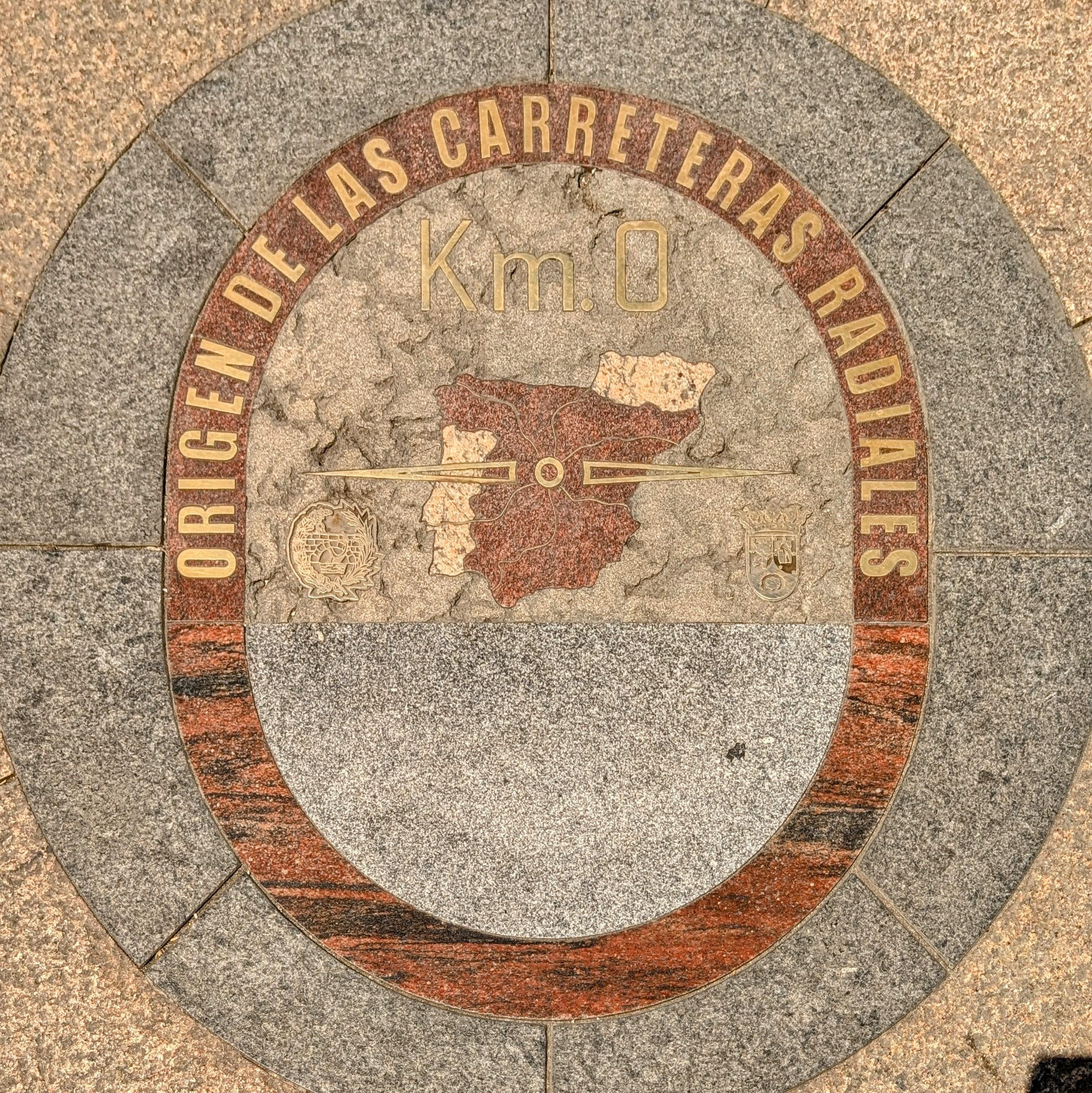 Circular plaque embedded in the ground marked "Origen de las Carreteras Radiales" with "Km. 0" at the center. Features a map of Spain with radial lines and a coat of arms. Made from stone, with an outer reddish-brown ring and a central grey area.