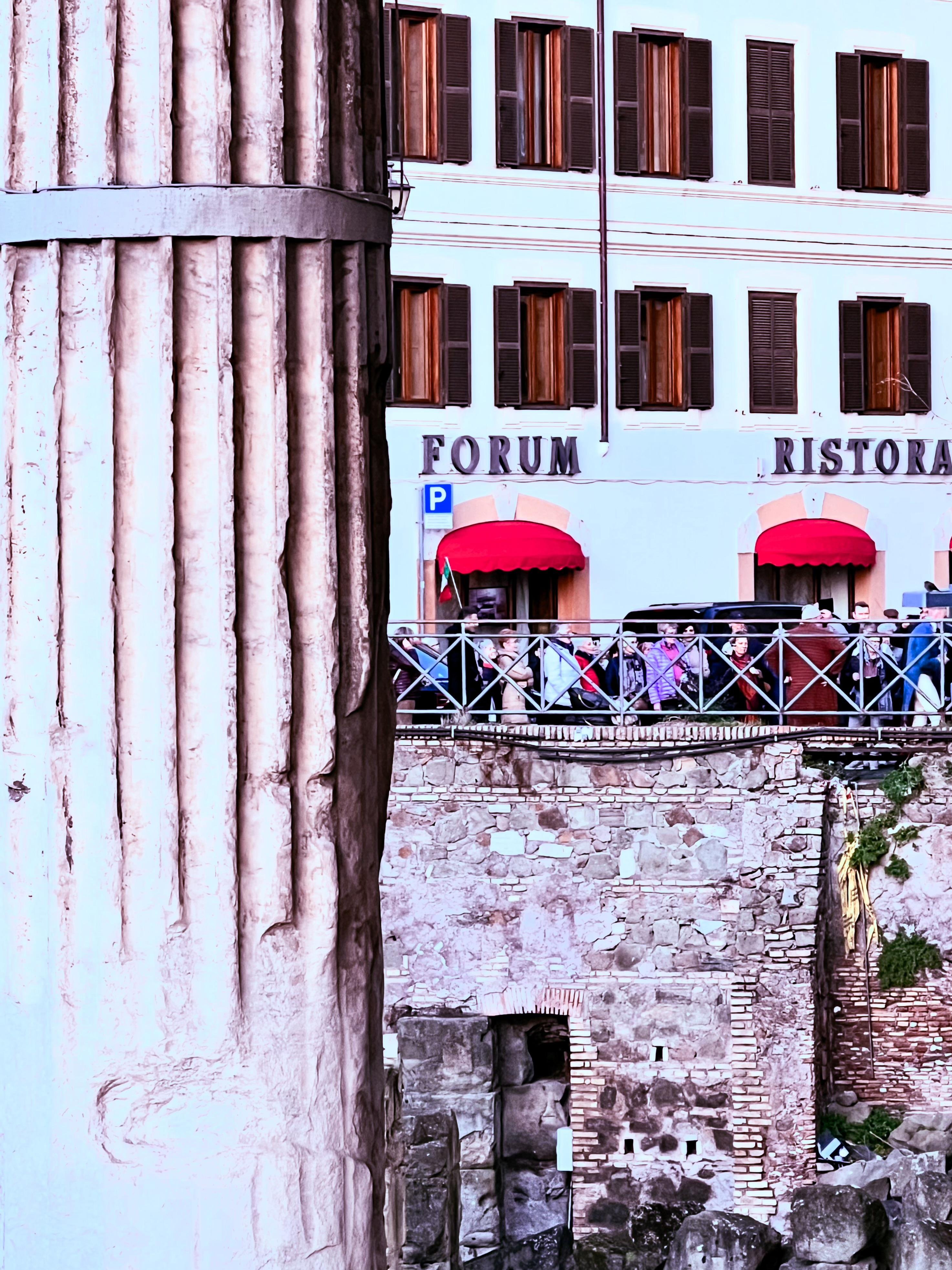 Grande colonna antica sulla sinistra, che sembra parte di una rovina storica. Sullo sfondo, c'è un edificio con un'insegna che dice "FORUM RISTORANTE". L'edificio ha diverse finestre con persiane e tende rosse sopra le entrate. Ci sono anche muri di pietra e un po' di vegetazione, suggerendo che l'ambientazione possa avere un'importanza storica.