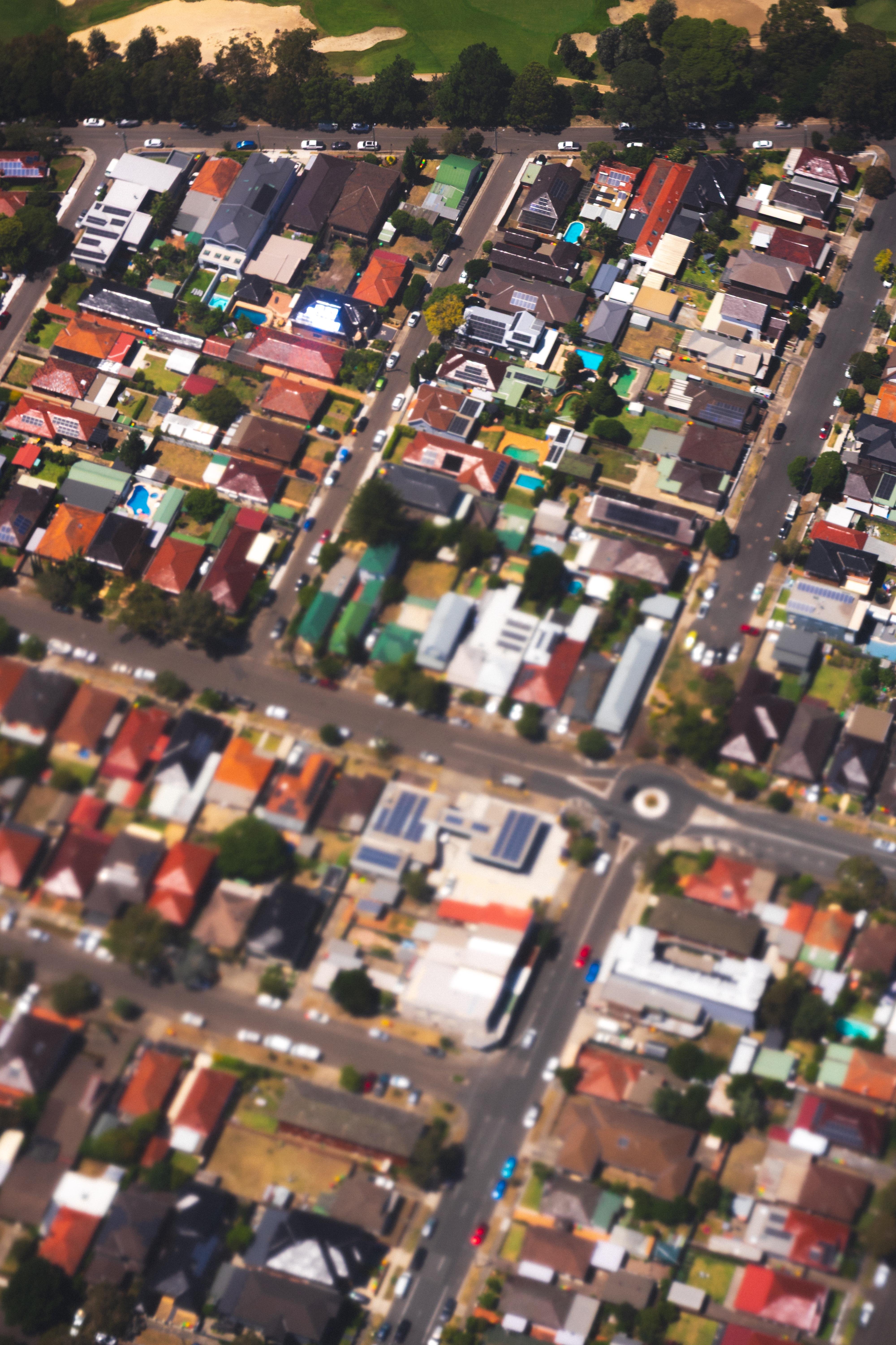 Aerial view of a suburban neighborhood with rows of houses aligned along parallel streets. Houses have varied roof colors, including red, gray, and blue. A central roundabout is visible with streets branching out. Sparse greenery is scattered throughout, and a golf course with sand traps and green patches is visible at the top of the image.