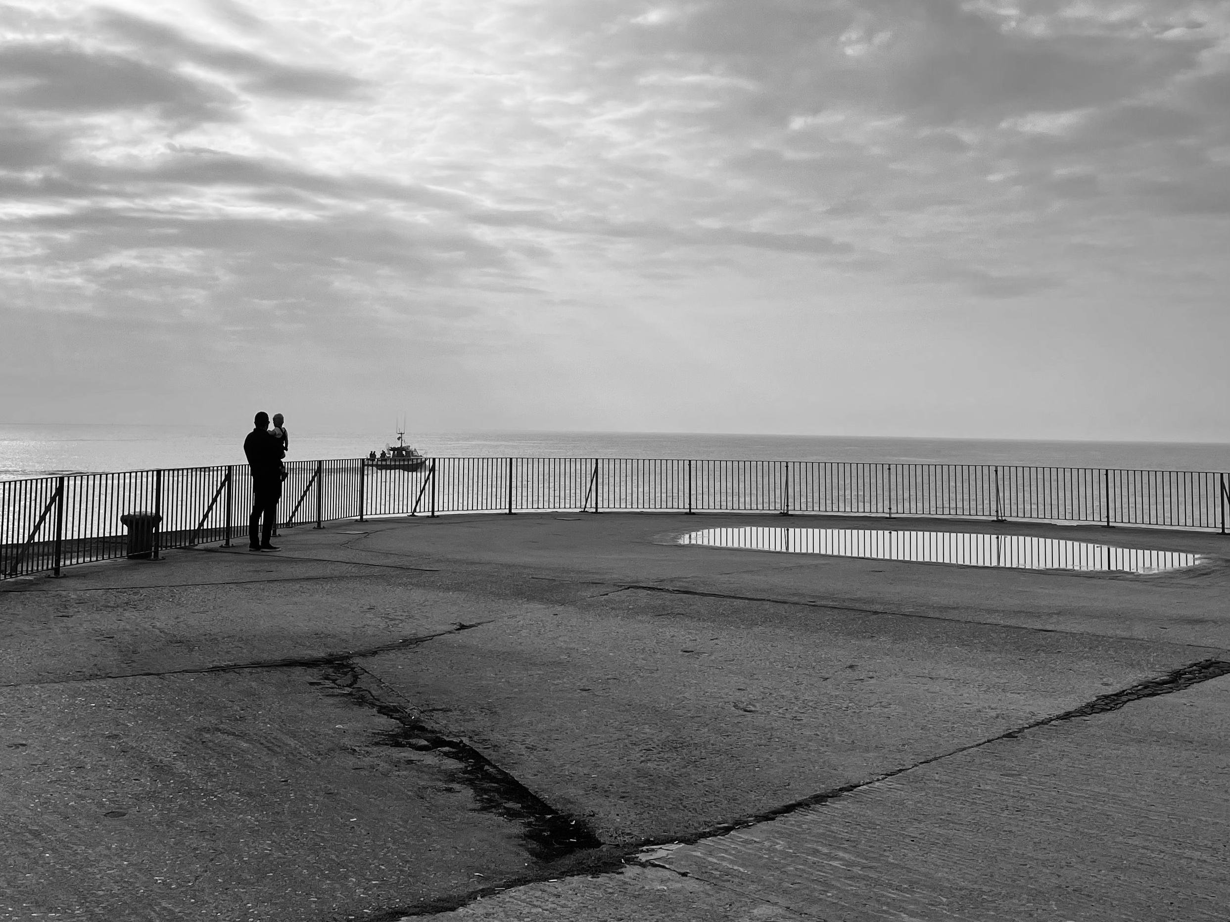 The image shows a black and white scene of a man holding a child standing near a railing, looking out at the sea. A boat is visible in the distance on the water. The ground is paved, and there's a puddle reflecting the sky. The atmosphere is calm, with a partly cloudy sky.