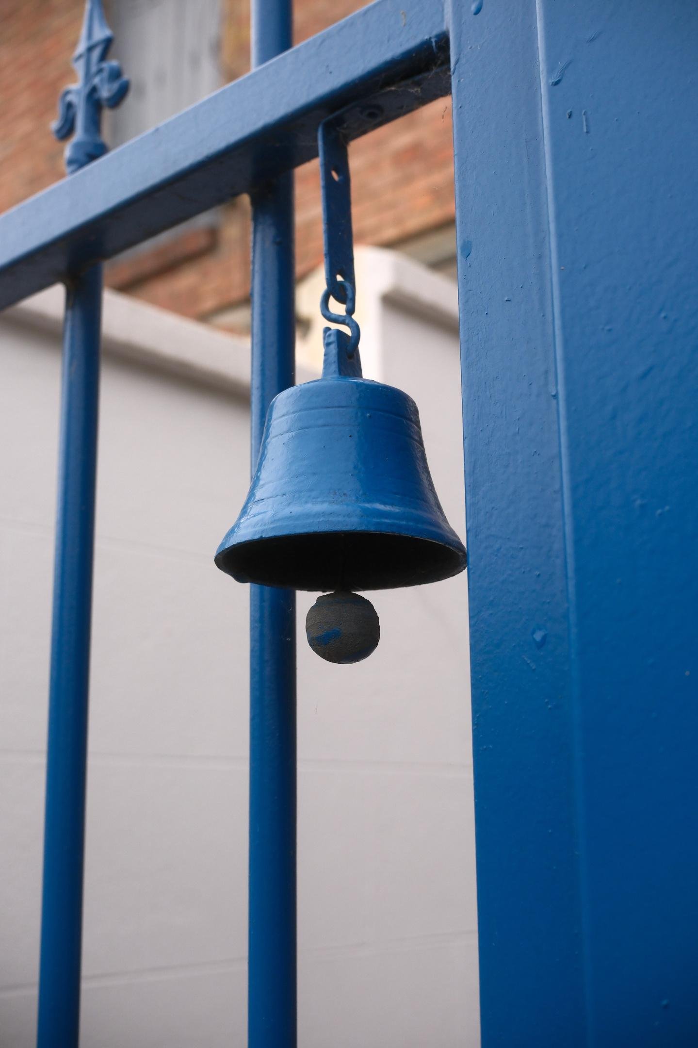 Photo verticale. Gros plan d’une cloche bleu pendue à un portail en fer peint dans le même bleu