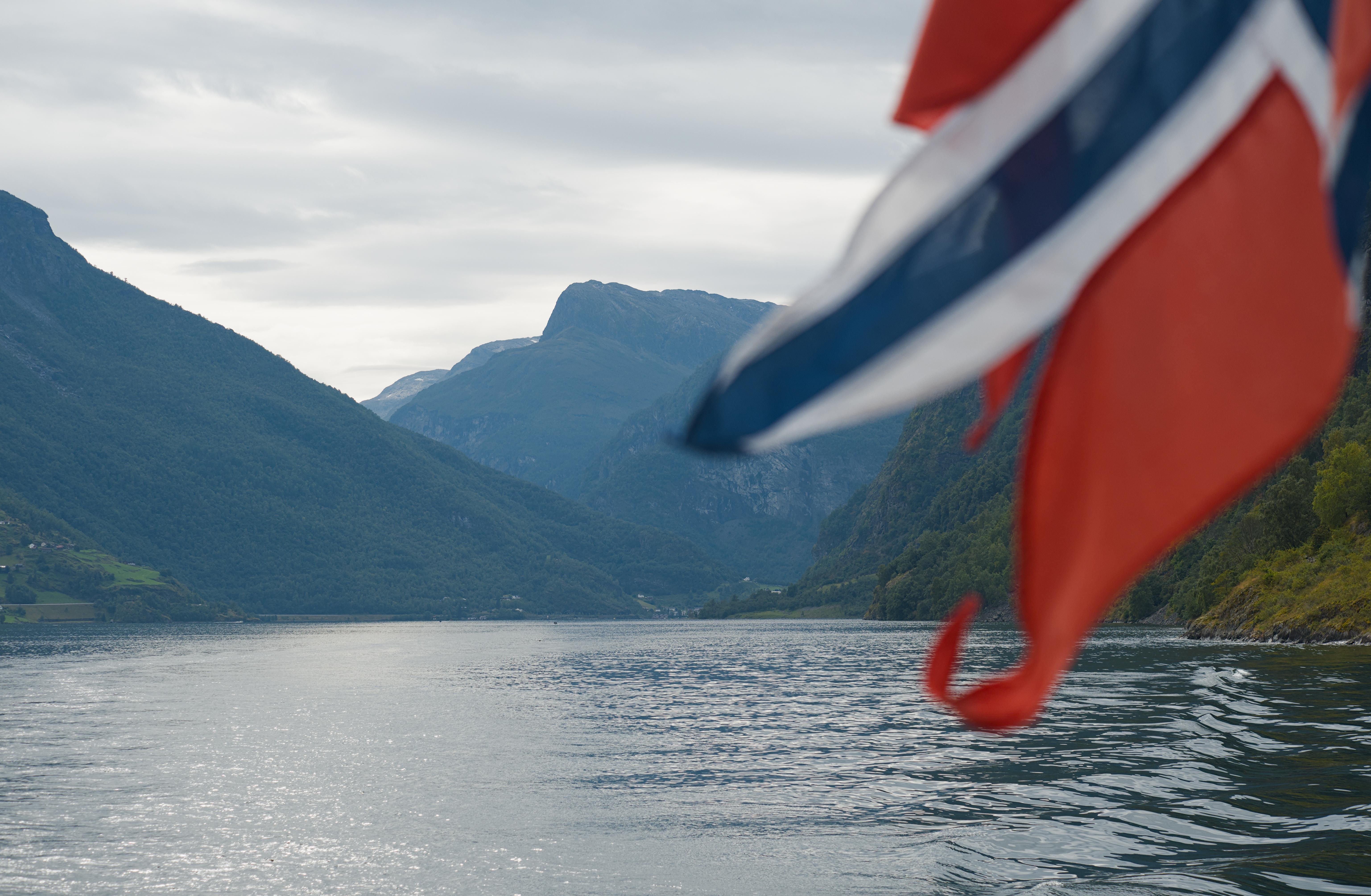 Norwegian flag in the right foreground, partially blurred, overlooking a calm fjord with steep green mountains on both sides under a cloudy sky.