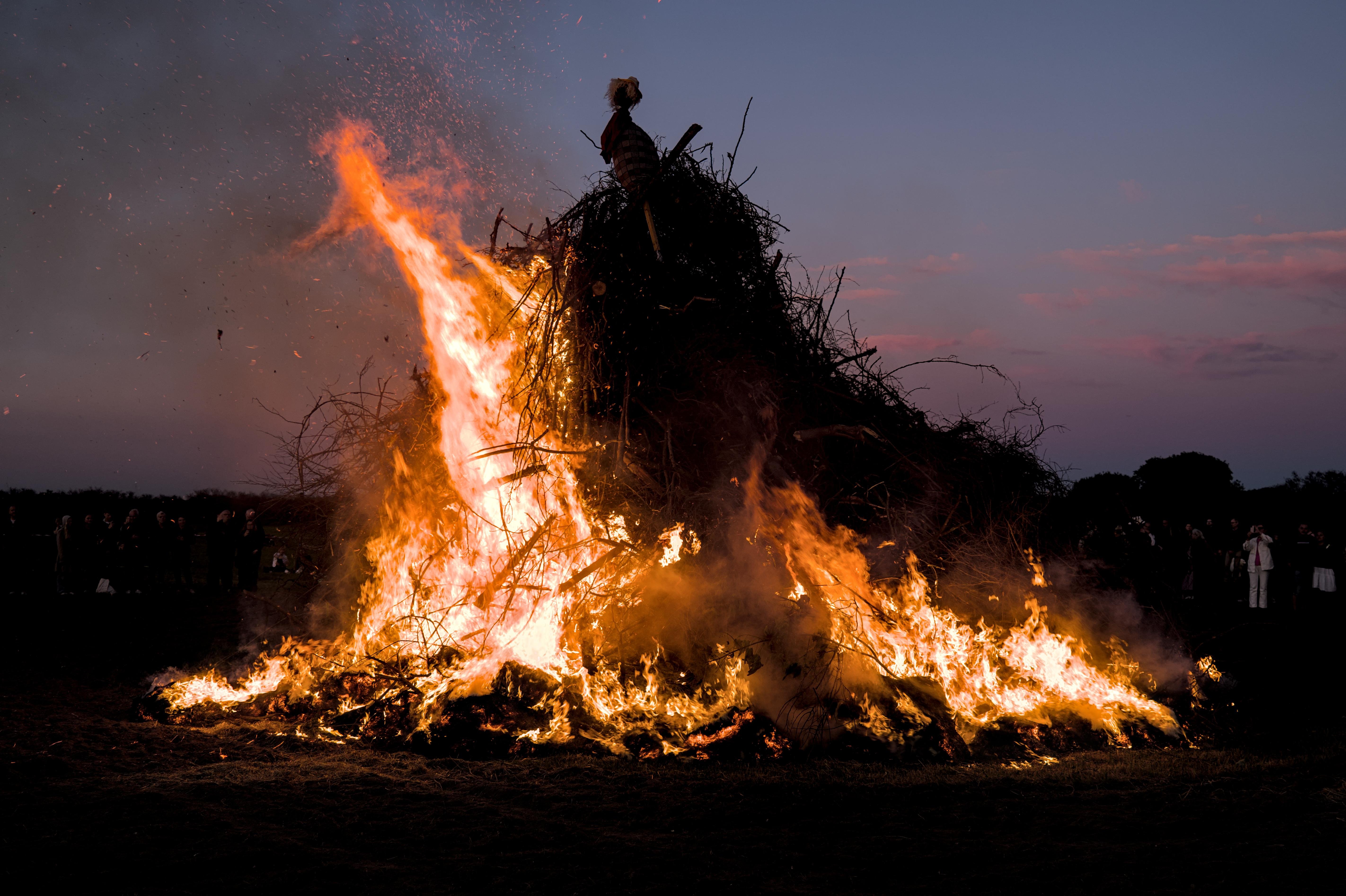 This image shows a large bonfire at dusk or nighttime. It appears to be part of a traditional or cultural event, as there are people in the background watching the flames. The fire is quite intense, with tall flames and a significant amount of burning material, possibly wood and brush.