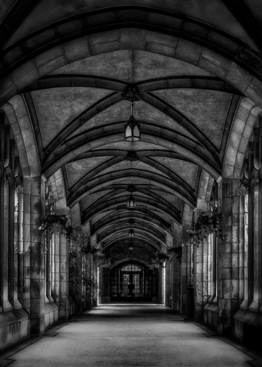 Black-and-white view down a long stone corridor with repeating Gothic arches, ribbed vaulted ceiling, hanging lanterns, and tall side windows leading to a glass-paned door at the far end.