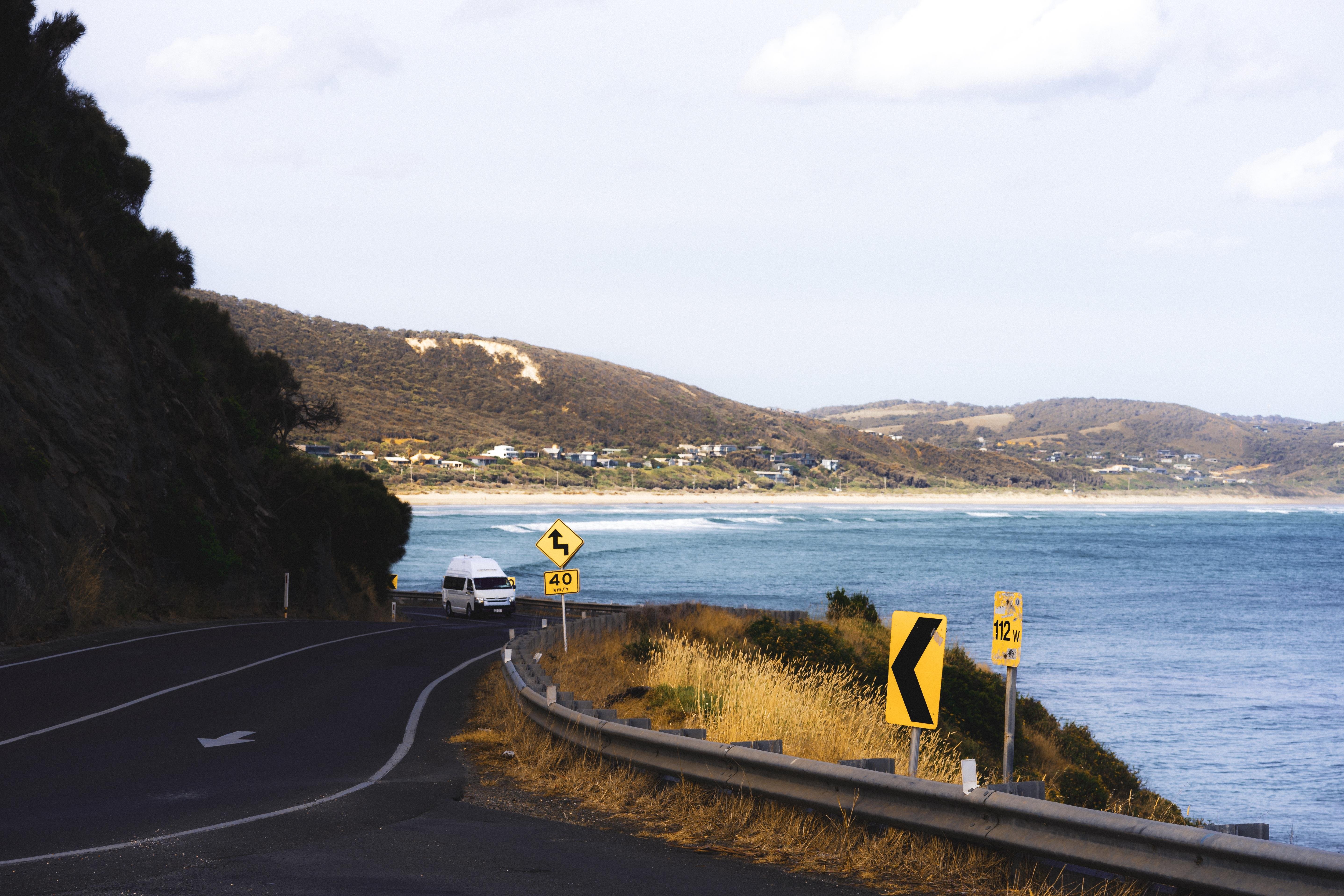 The image shows a coastal road with a vehicle driving along it. The road has a curve and is lined with guardrails. There are several road signs indicating a sharp turn and a speed limit of 40. To the right, there is an expanse of water, likely the ocean, and in the background, there are hills with scattered houses. The setting appears to be a scenic coastal area.