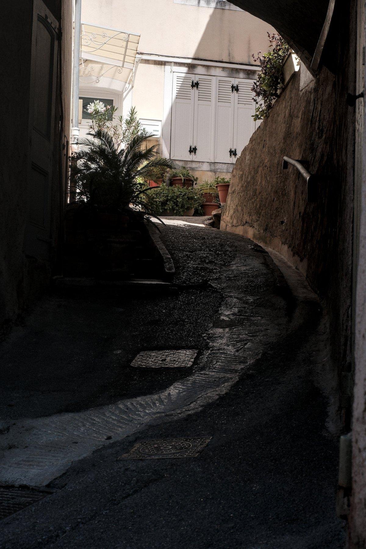 Photo verticale. Un passage en pierre dans l’ombre menant à une porte d’entrée de maison. une double fenêtre aux volets blancs fermés est à droite au bout du passage. Des pots de fleurs avec des plantes vertes sont en dessous de la fenêtre. La porte d’entrée est abritée par un petit toit en verre poli