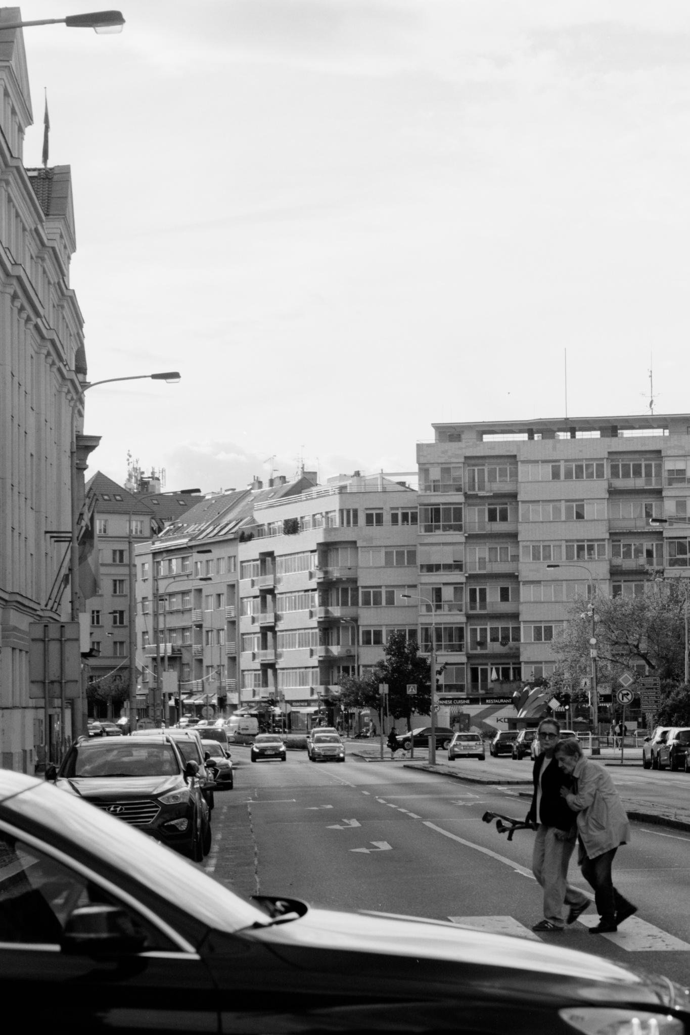 Black-and-white street scene with mid-rise apartment buildings in the background, parked cars along the left curb, and moving traffic on a multi-lane road; two pedestrians cross at a crosswalk in the lower right while a car passes in the foreground.