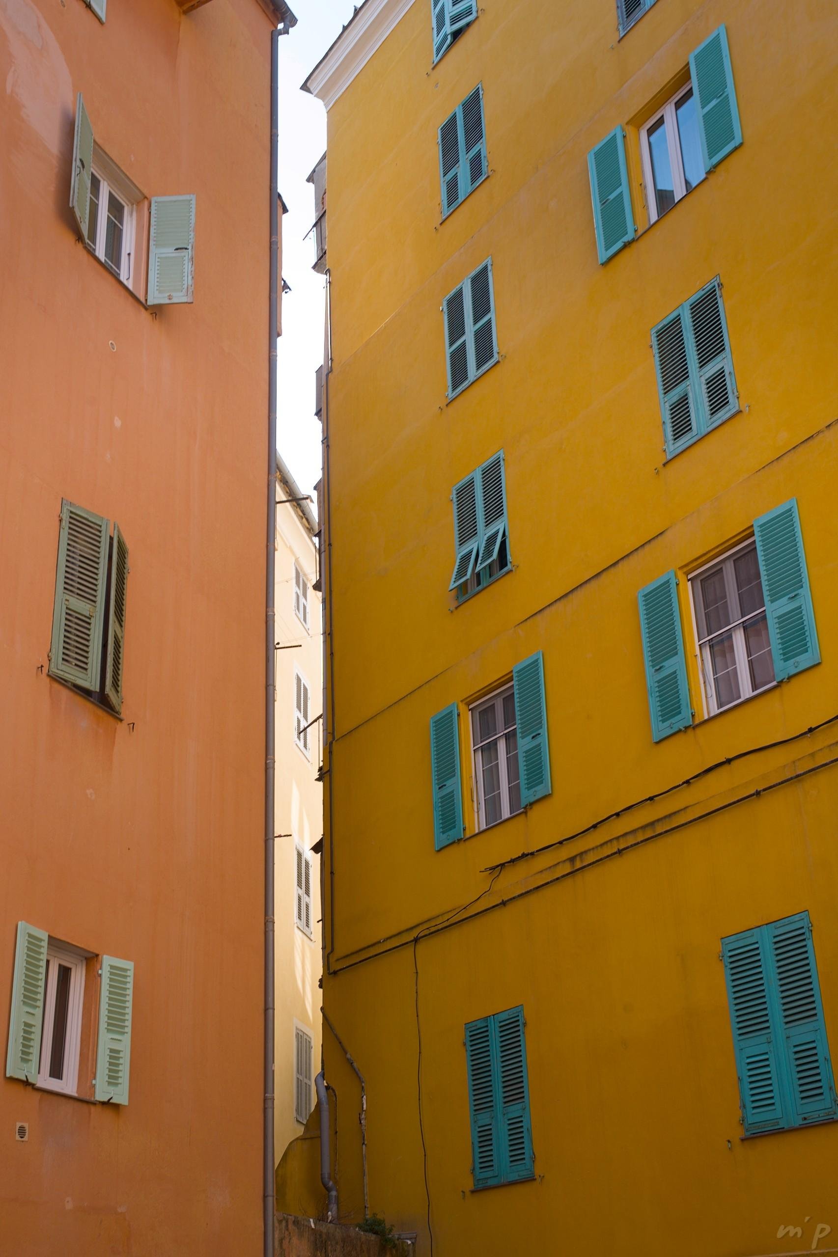 🇬🇧 Vertical color photo of two tall buildings at right angles with a small space in between. On the left the facade is orange pierced with windows with sea green shutters, on the right the facade is yellow pierced with windows with emerald blue shutters. Through the space in between, another ocher building facade with gray shutters.

///


🇫🇷 Photo couleur verticale de deux hauts bâtiments à angle droit avec un petit espace entre les deux. À gauche la façade est orange percée de fenêtres aux volets vert d'eau, à droite la façade est jaune percée de fenêtre aux volets bleu émeraude. À travers l'espace entre les deux, une autre façade de bâtiment ocre avec des volets gris.