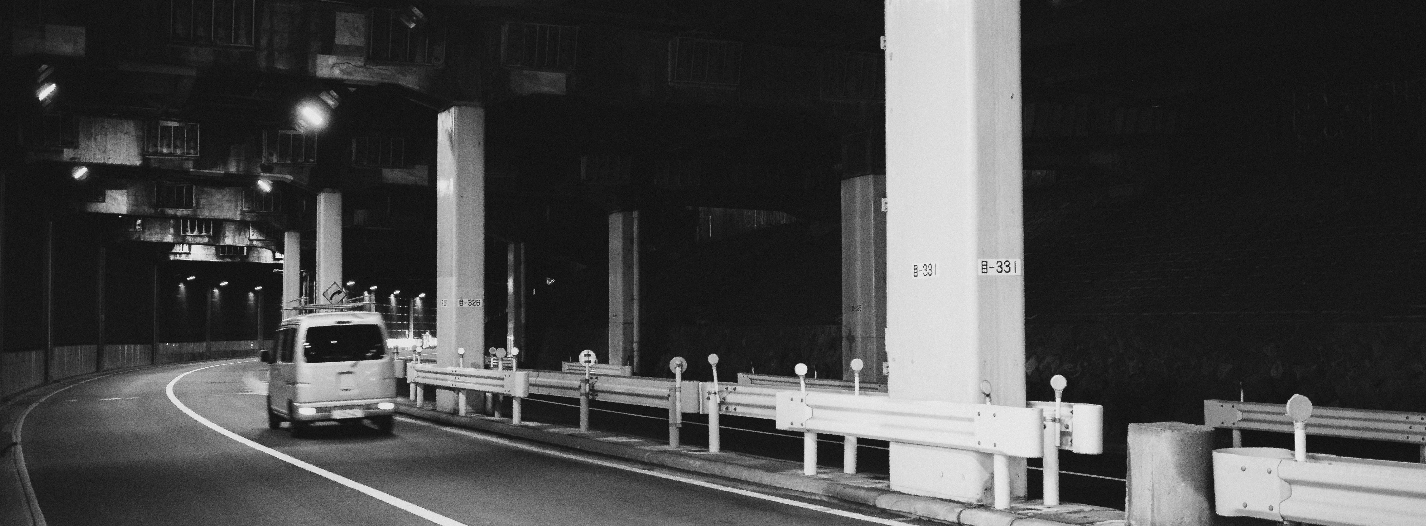 The image depicts a black and white scene of a van driving through a tunnel or underpass. The road curves to the left, and there are large concrete pillars supporting the structure above. The area is illuminated by overhead lighting, creating a stark contrast with the shadows in the tunnel. There are also guardrails alongside the road.