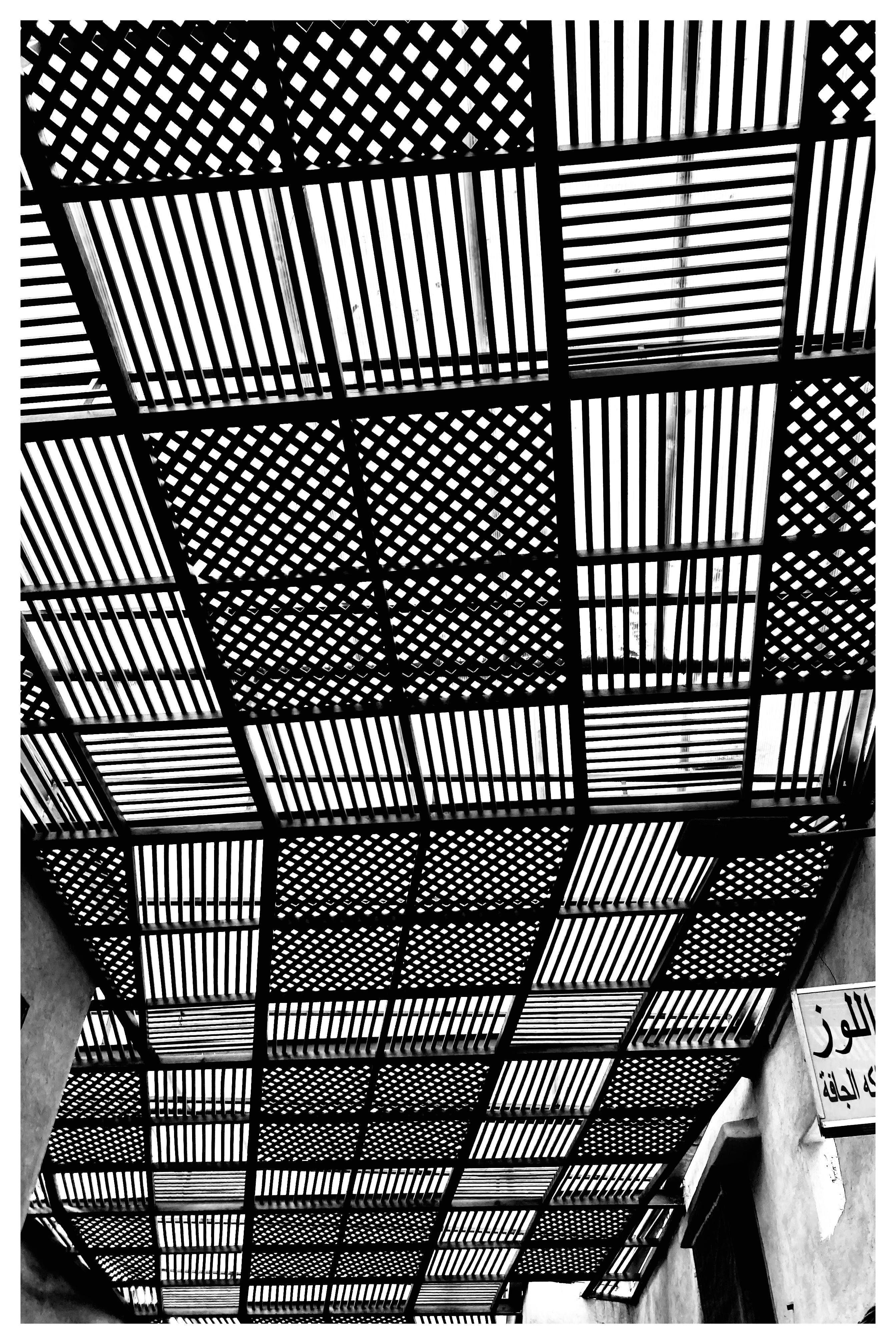 Black and white photo of a lattice-style ceiling composed of alternating diagonal and straight patterns, casting complex shadows. A sign with Arabic script is partially visible on the right side, attached to a wall below the ceiling.