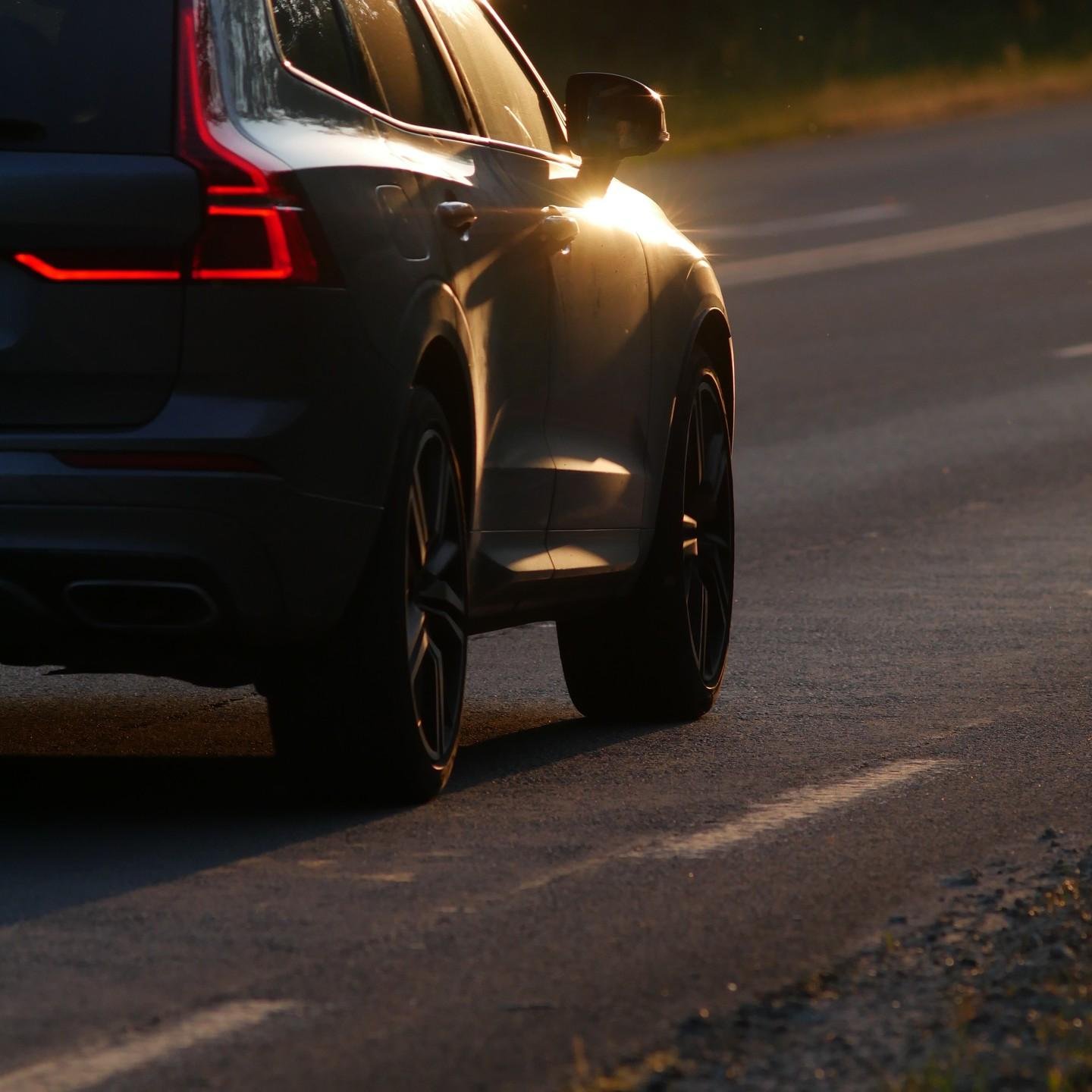 The image shows the rear side of a dark-colored car driving on a road during sunset. The car has illuminated rear lights, and the road has markings. The lighting suggests a warm or golden hour setting.