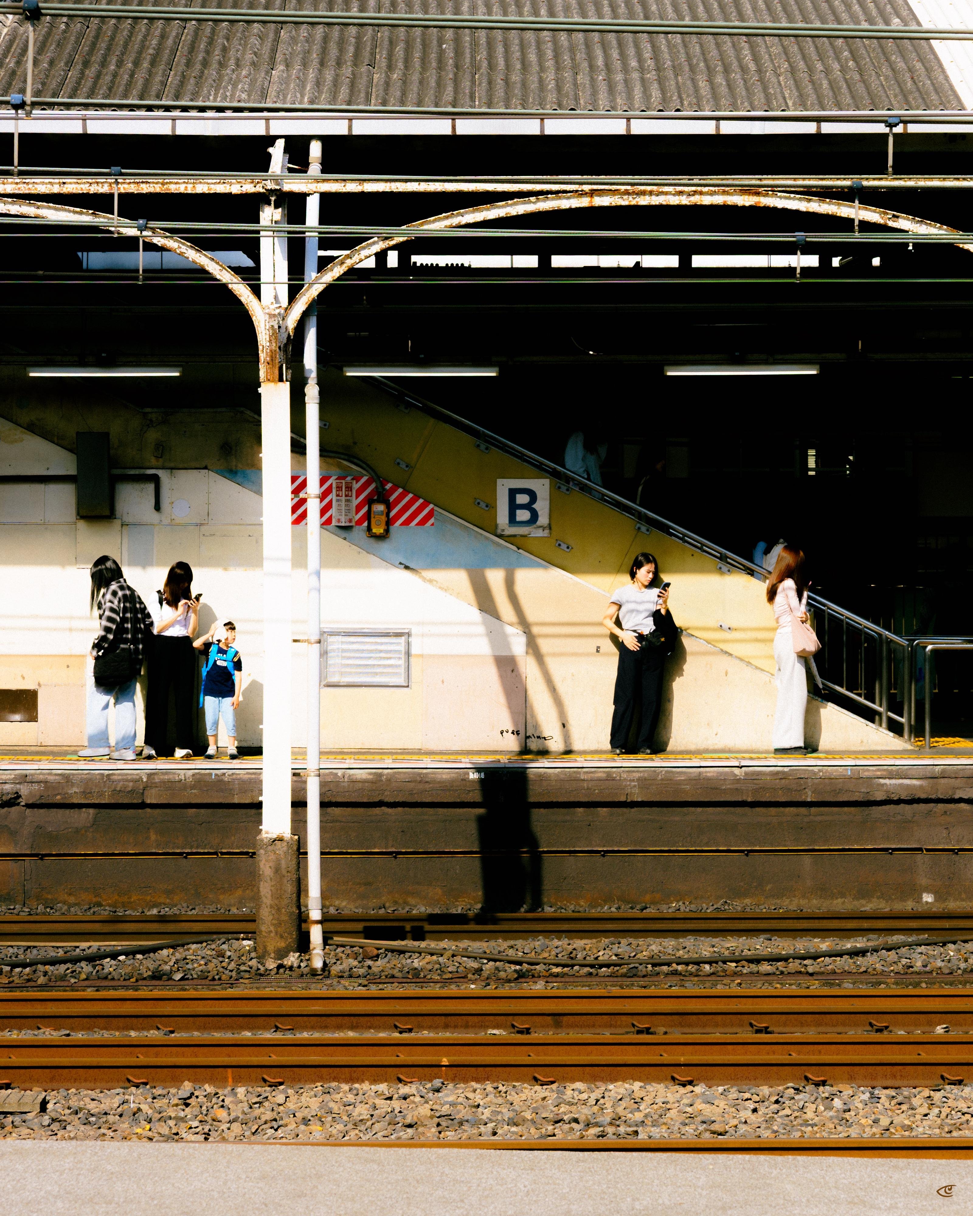 Train station platform with several people waiting, a child in a blue outfit, and two women looking at phones; overhead metal canopy and columns, a stairway marked with a large “B,” and train tracks in the foreground with long shadows cast across the platform.