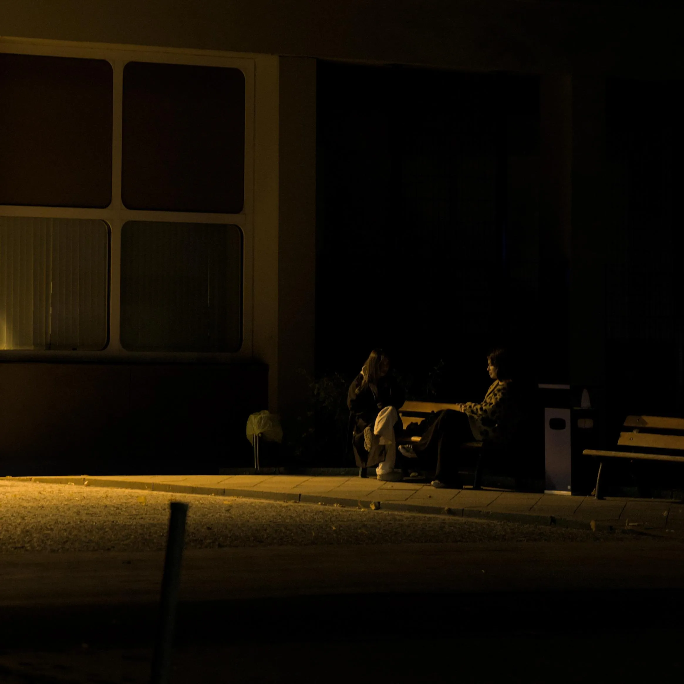 Photograph of two women sitting on the same bench turning to face each other at nighttime, apparently in the middle of a conversation, in front of a building with a large window with closed interior blinds. Most of the image is very dark and the only light is a yellow light comes from an unseen source to the left of the image. Each of the two persons on the bench is only illuminated by a thin ray of this yellow light on one of their sides in a chiaroscuro effect.