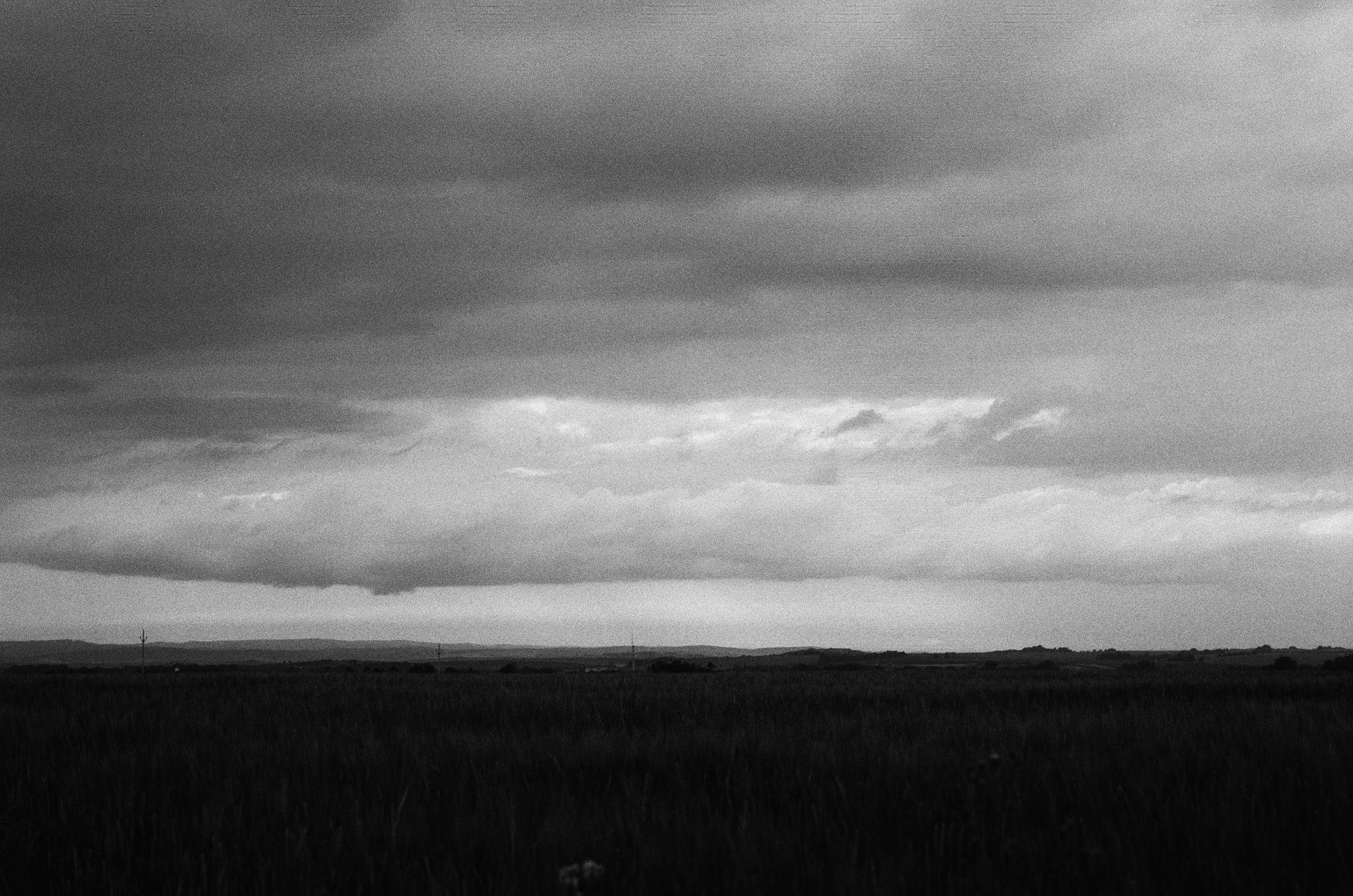 Black-and-white landscape with a dark grassy field in the foreground, a low flat horizon in the distance, and layered storm clouds filling most of the sky.