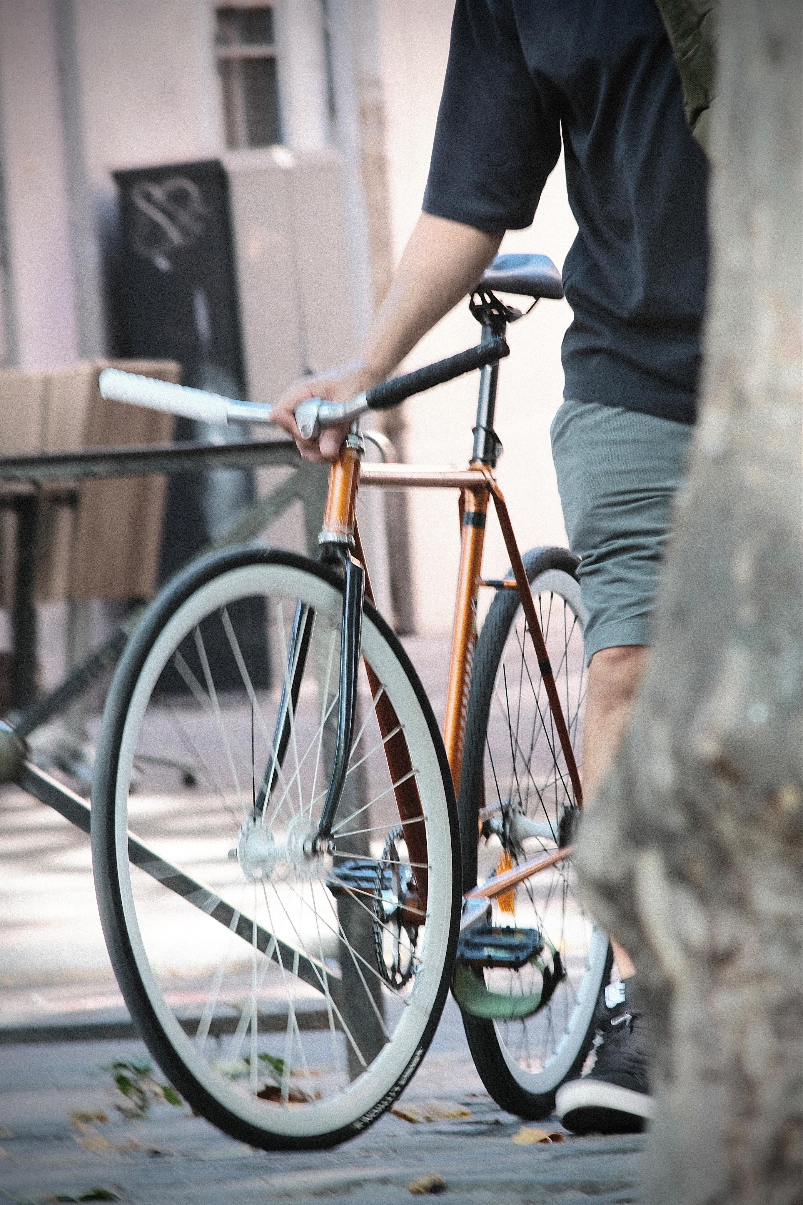 Person walking and holding a copper-colored single-speed bicycle with white wheels on a city sidewalk; torso and legs visible with a tree trunk blurred in the foreground.