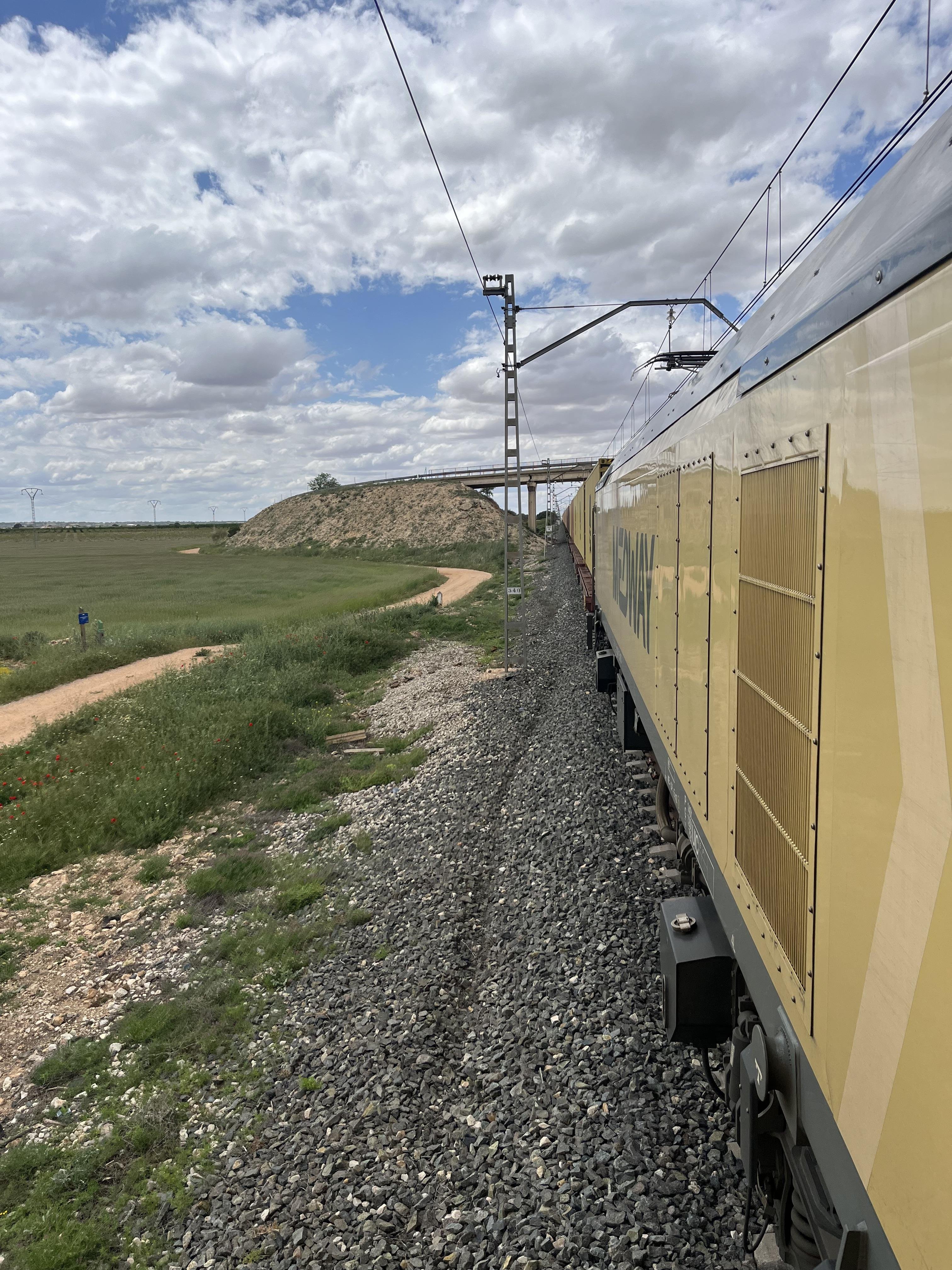 The image shows a train traveling along a track in a rural area. The landscape features a rocky railway line beside grassy fields and a dirt path. There is an overhead bridge in the distance. The sky is partly cloudy, with sunlight filtering through.