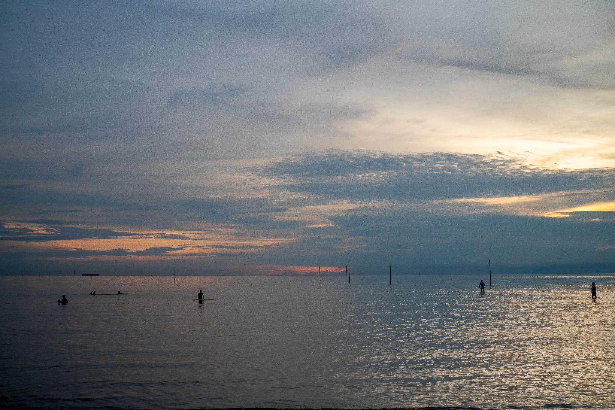 "Serene seascape during sunrise or sunset with a calm ocean stretching to the horizon. The sky is filled with soft clouds illuminated in orange and blue tones. Several people are in the water, standing or paddleboarding, while thin poles protrude from the sea, possibly marking boundaries. The tranquil light and gentle waves create a peaceful, scenic atmosphere