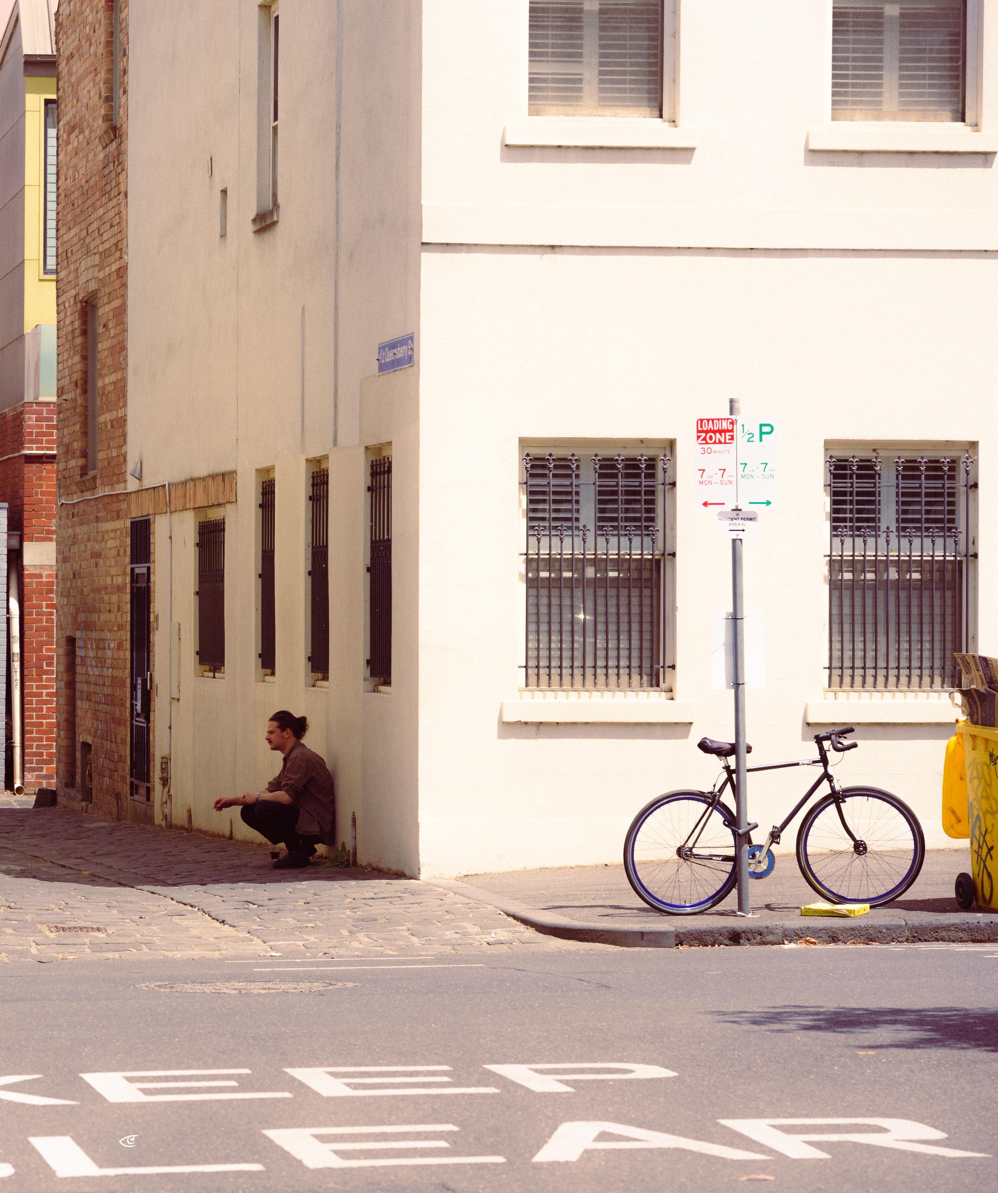 Man crouching on a cobblestone lane beside a cream-colored building with barred windows; a black bicycle is locked to a street sign pole on the sidewalk, with “KEEP CLEAR” painted on the road in the foreground.