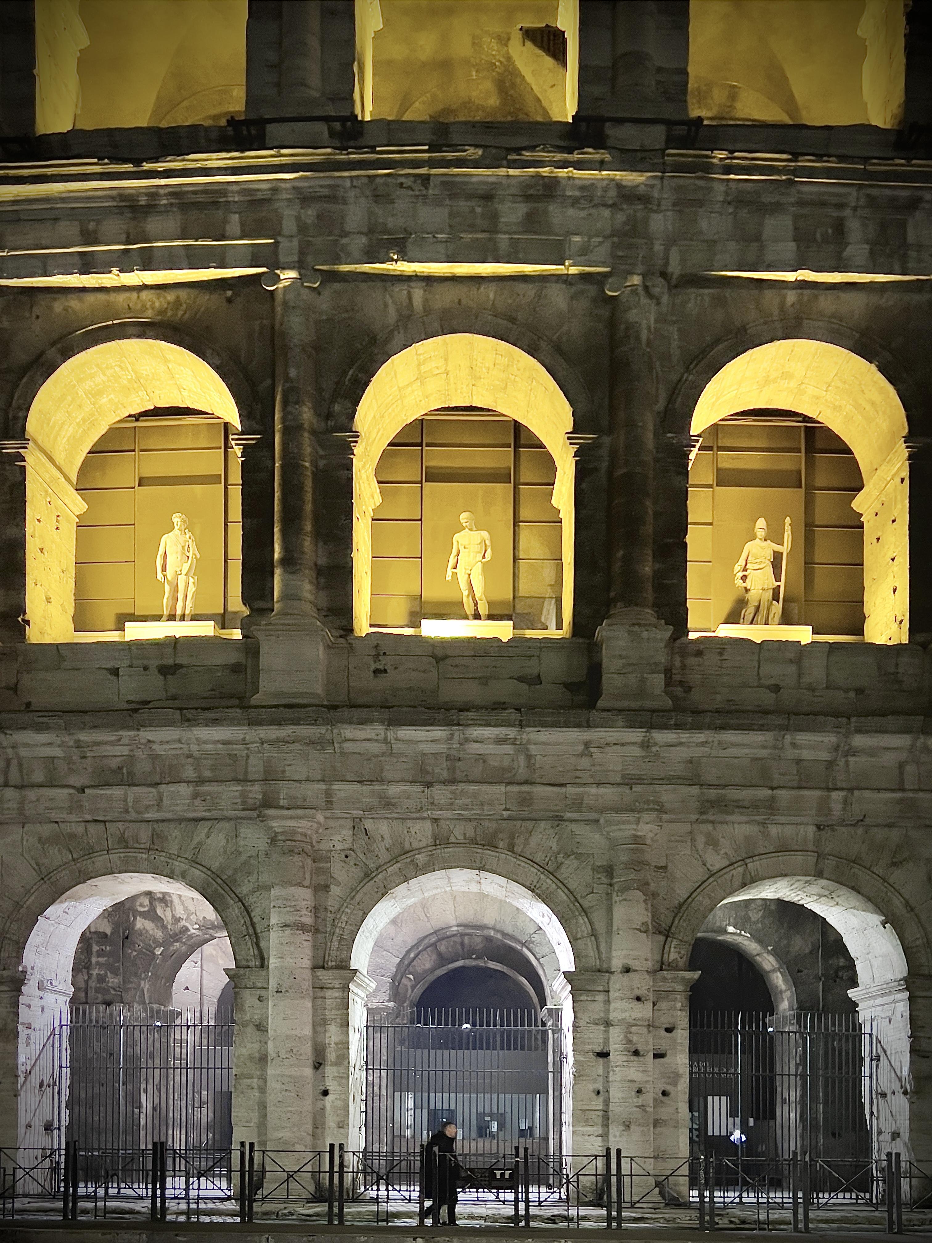 Primo piano notturno della facciata in pietra del Colosseo con due ordini di arcate sovrapposte; tre statue illuminate occupano gli archi superiori, mentre quelli inferiori sono chiusi da cancellate metalliche con un passante che cammina lungo la strada antistante.