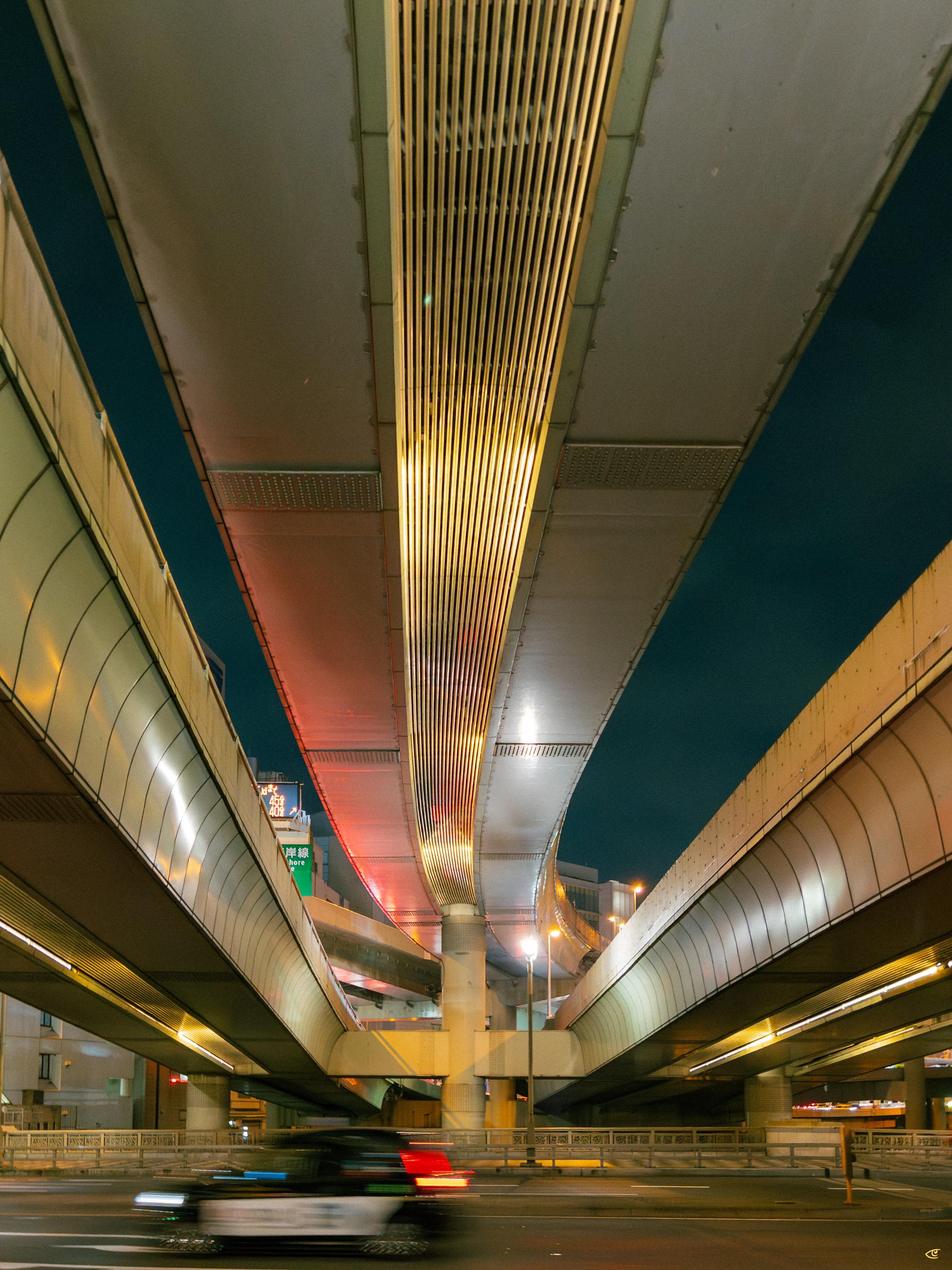 View from street level beneath intersecting elevated highways at night, with a large central vented panel and steel beams overhead, concrete pillars supporting the overpasses, and a blurred car passing along the road in the foreground.