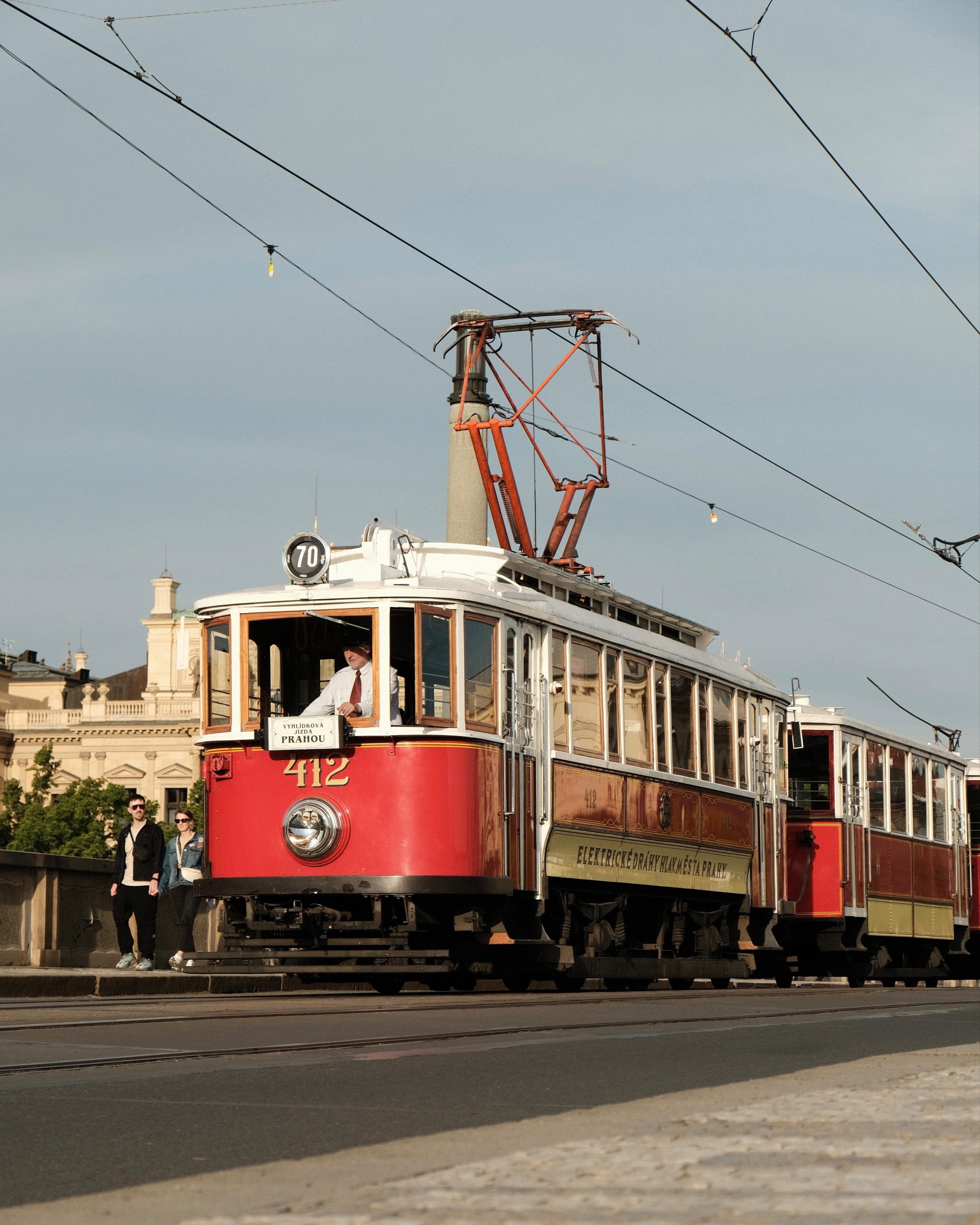 Red and cream vintage tramcar numbered 412 traveling on a city street under overhead power lines, with two pedestrians on the sidewalk and historic buildings in the background.