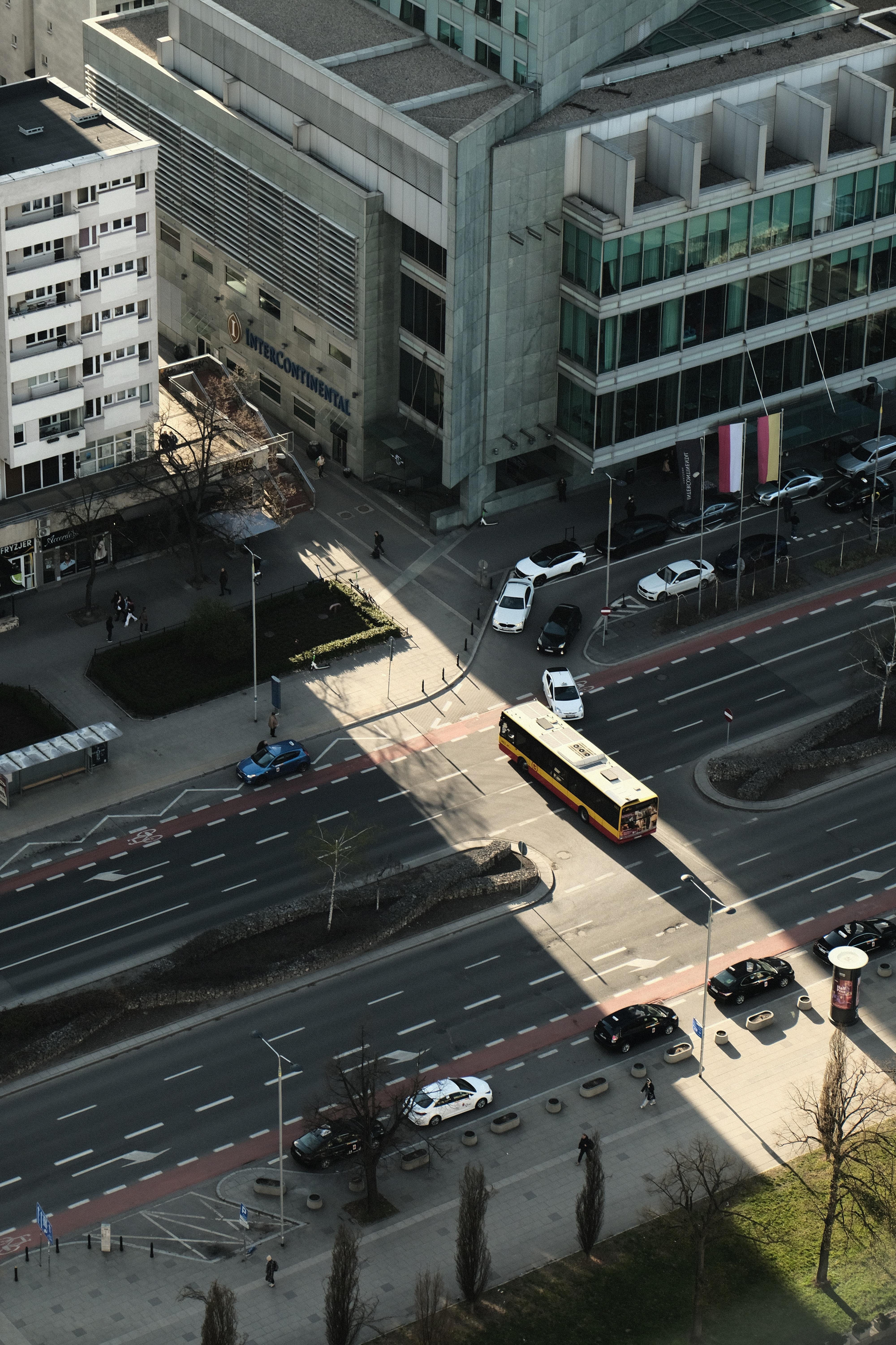 View of a street crossing from an angle above. Tiny cars driving on the roads. The low afternoon sun shines along one of the streets leading to the crossing, the sun lights a narrow band across the crossing, contrasting the surrounding shadows of the city buildings. Within the light band, a bus is taking a turn on the crossing. This literally highlights the bus against the other cars.
