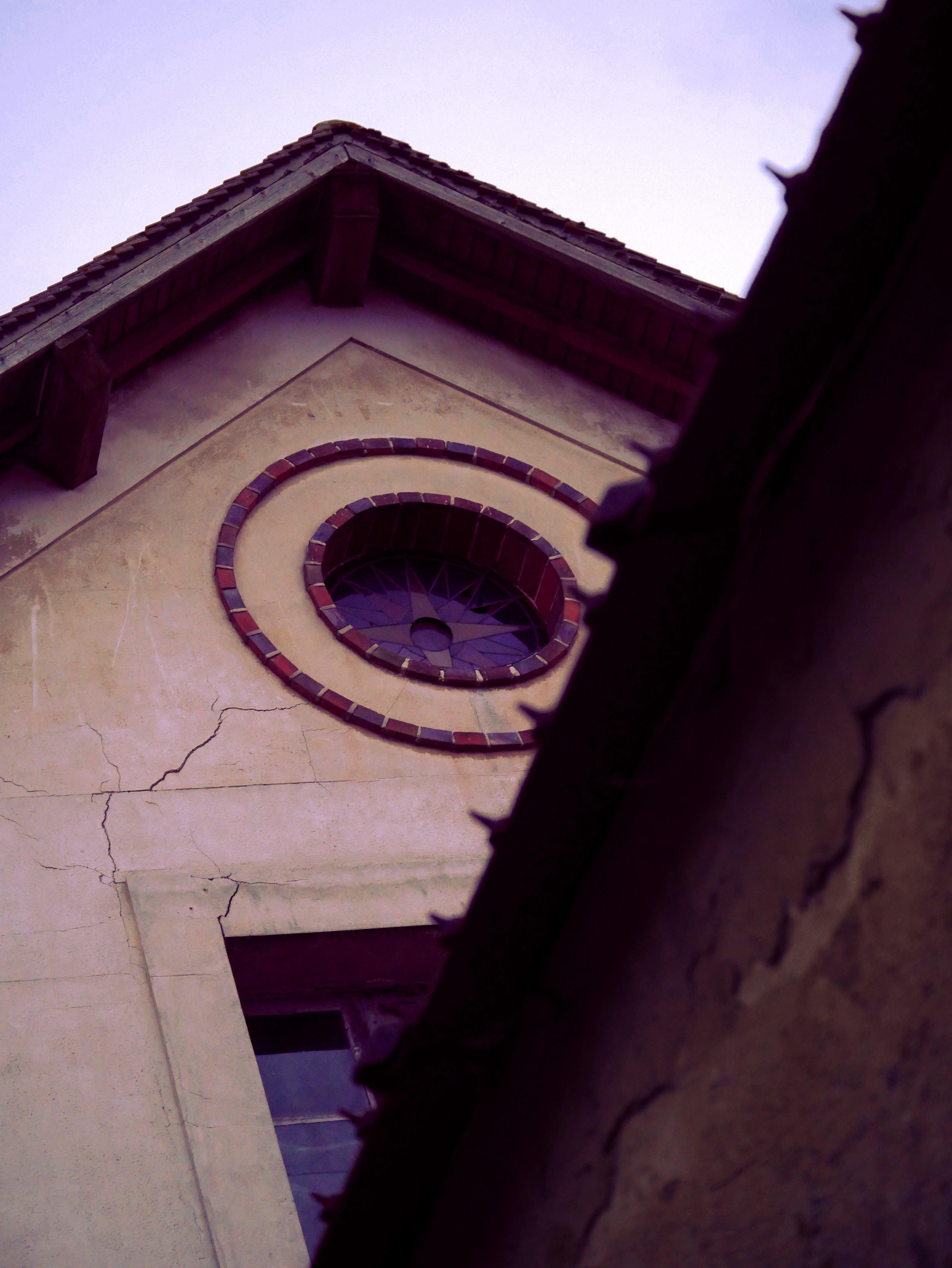 The image shows an architectural detail of a building, specifically the top part of a facade. It features a circular stained glass window set within a larger triangular gable, with visible cracks in the plaster or concrete surface. The roof is sloped with shingles, and the scene has a slightly worn, vintage look.