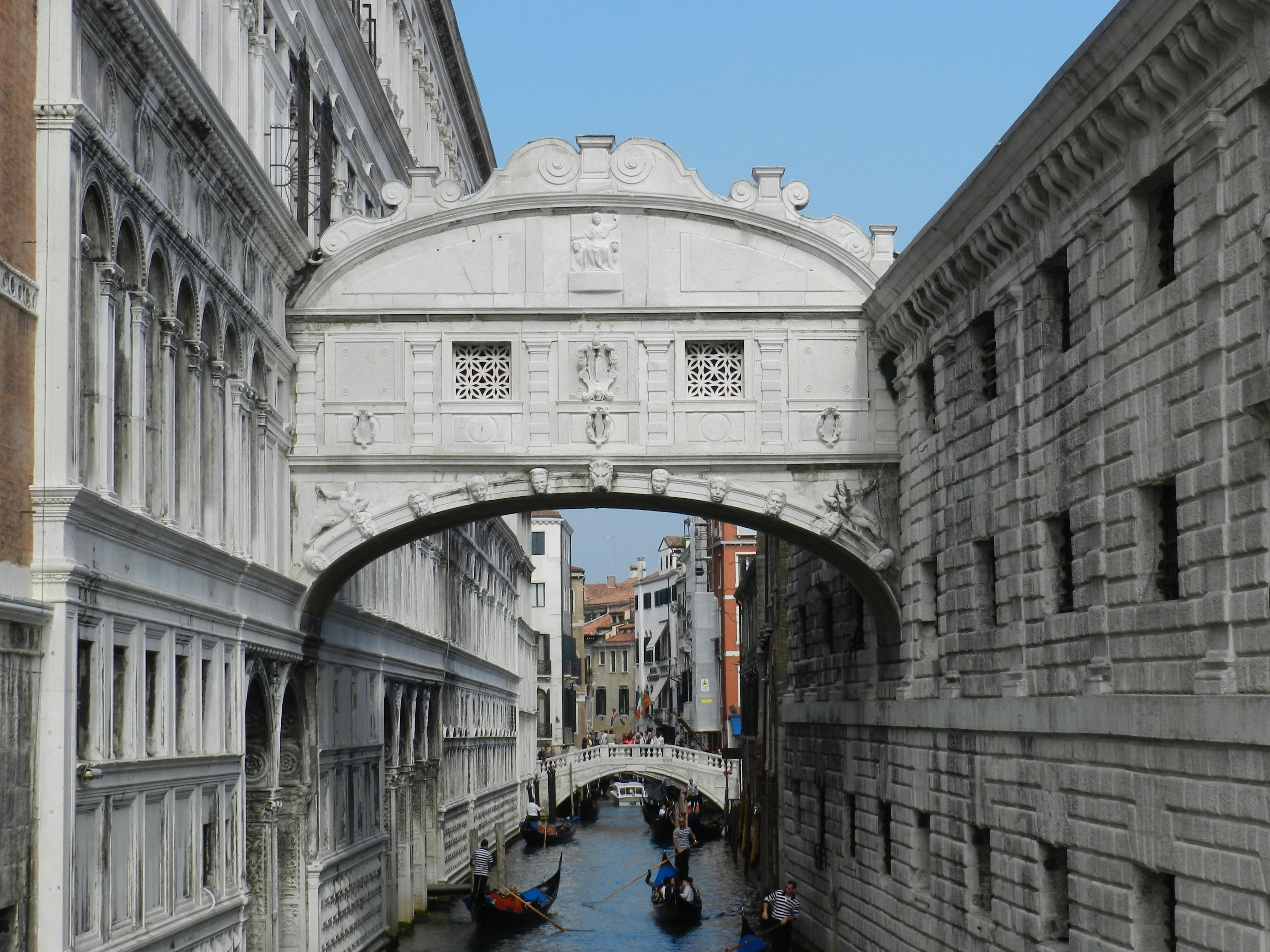 Photograph of the Ponte dei Sospiri (Bridge of Sighs) in Venice, Italy, captured from a viewpoint that looks along the narrow canal beneath it. The enclosed white limestone bridge, built in 1600, arches gracefully between two historic buildings: the ornate façade of the Doge’s Palace on the left, with its elegant rows of arches and sculpted details, and the darker, fortress-like walls of the old prison on the right. The bridge features delicate stone lattice windows and low-relief carvings that add to its baroque character. Below, several gondolas glide through the canal, their gondoliers in traditional striped shirts steering tourists beneath this iconic landmark. In the background, the canal continues toward a smaller pedestrian bridge and pastel-colored Venetian buildings. The Bridge of Sighs famously connected the interrogation rooms of the Doge’s Palace to the prison—its poetic name inspired by the idea that prisoners would sigh at their final view of Venice through its small windows.