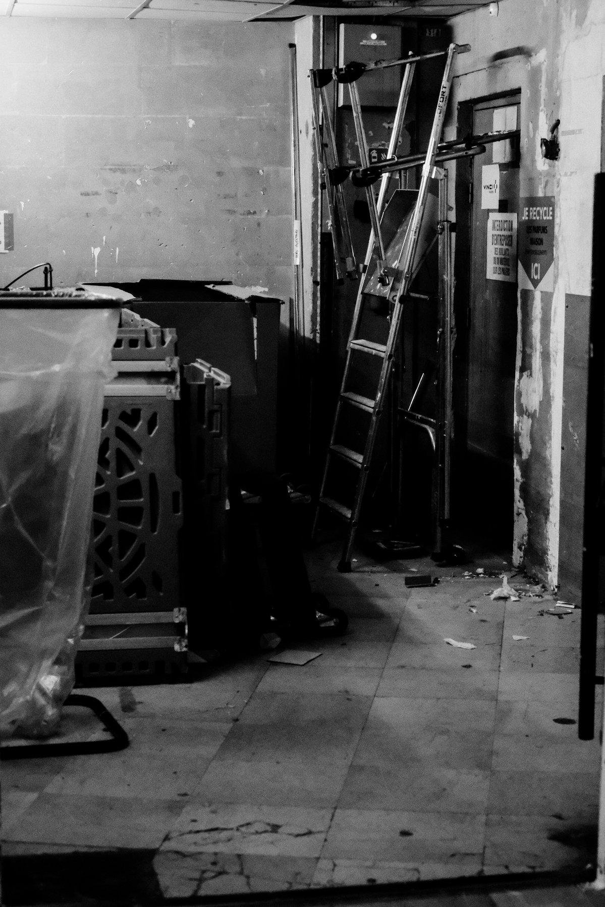 Photo verticale en noir et blanc. Un local en bazar, avec un escabeau, des panneaux en métal percés de motifs décoratifs, un cadre tenant un sac poubelle transparent, et une porte avec un sticker du logo Vinci collé