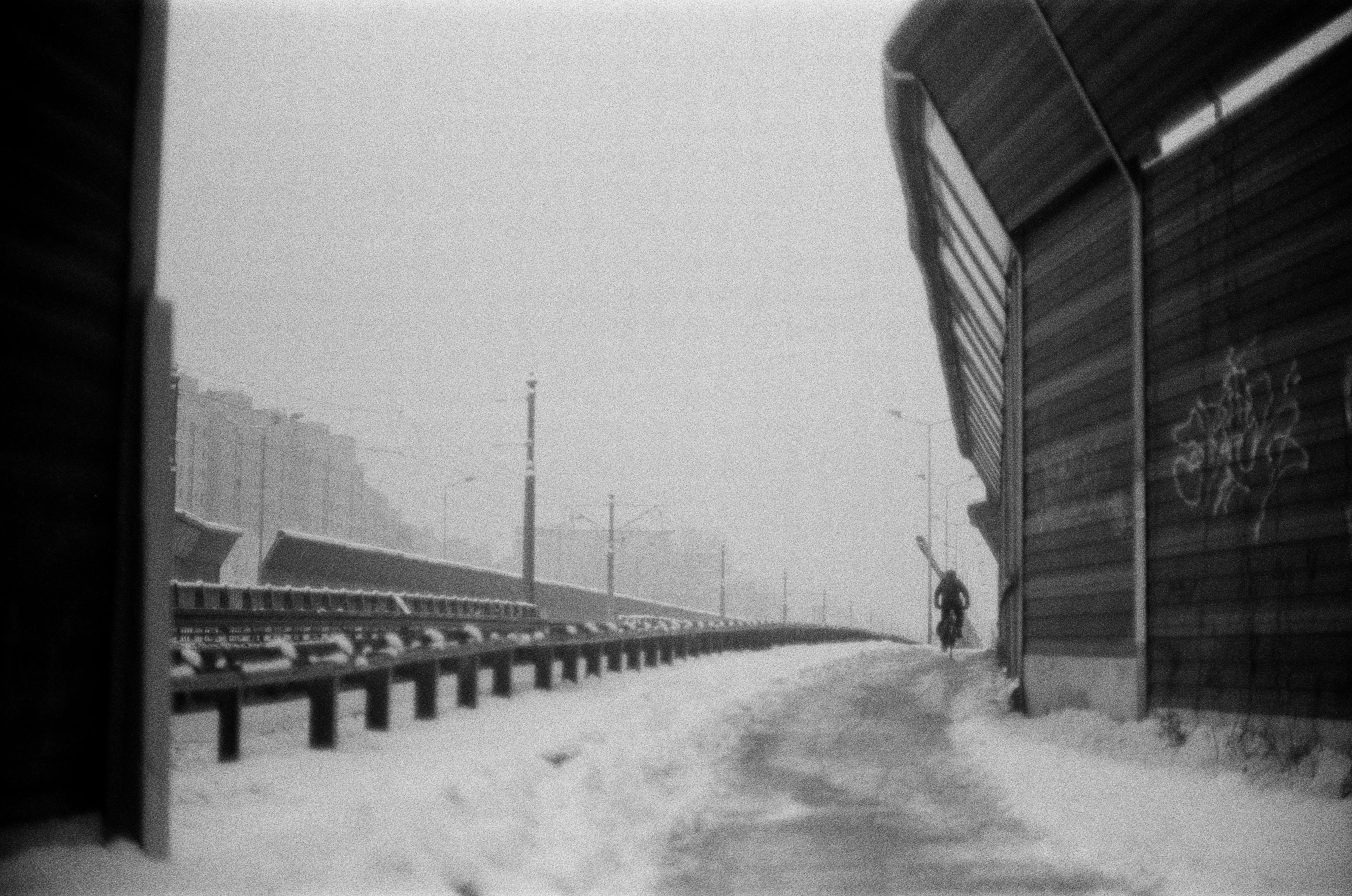 Black-and-white photo of a snow-covered road curving into the distance, with a guardrail and streetlights on the left, a tall sound barrier wall with graffiti on the right, and a lone cyclist riding away in the center-right under a hazy sky.