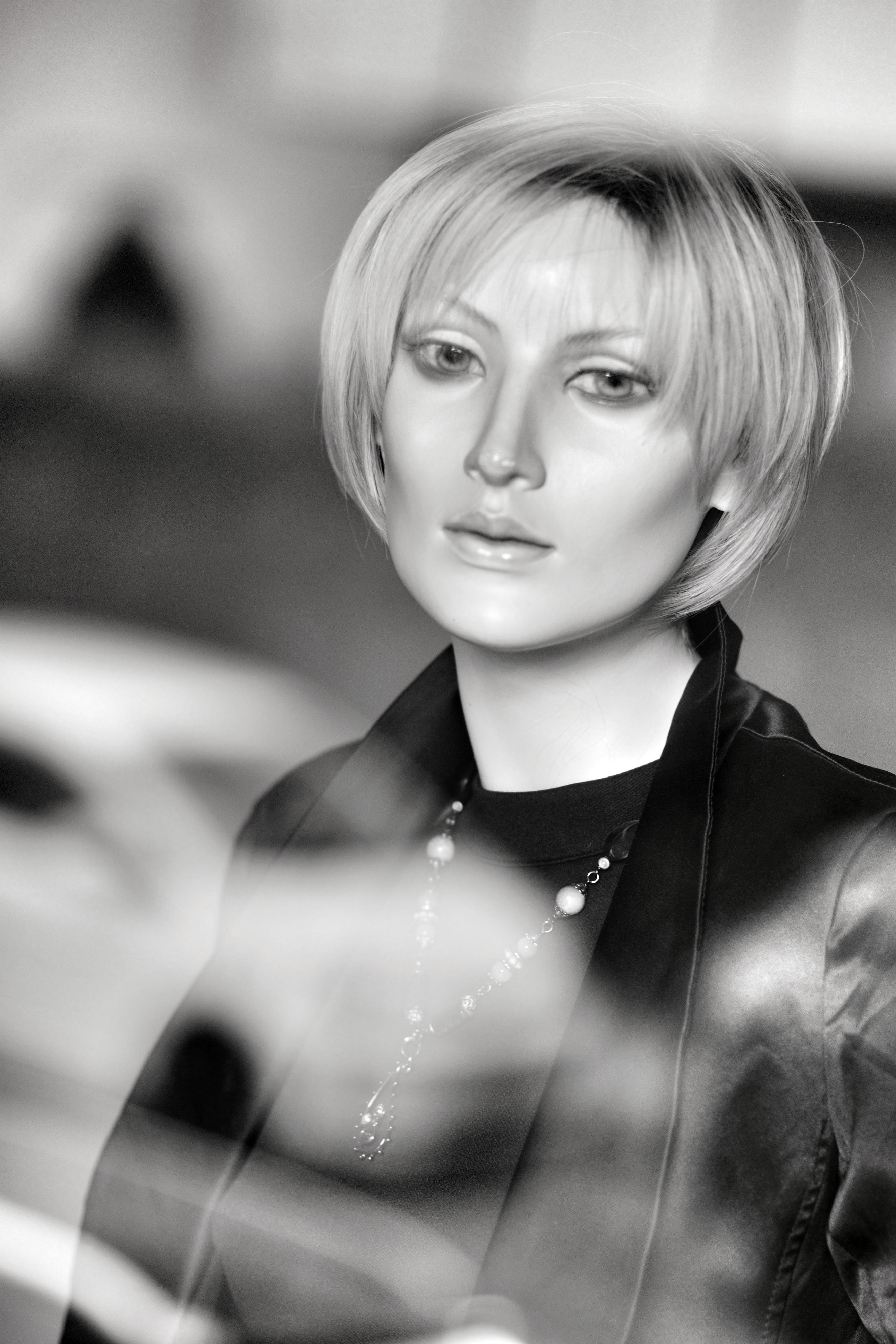 Black-and-white close-up portrait of a female mannequin with a short blonde bob haircut, wearing a dark jacket and necklace, facing the camera with a blurred background and foreground shapes.
###
Schwarz-weißes Nahaufnahme-Porträt einer weiblichen Schaufensterpuppe mit kurzem blondem Bob, die eine dunkle Jacke und eine Halskette trägt, der Kamera zugewandt ist und vor einem unscharfen Hintergrund sowie unscharfen Formen im Vordergrund steht.