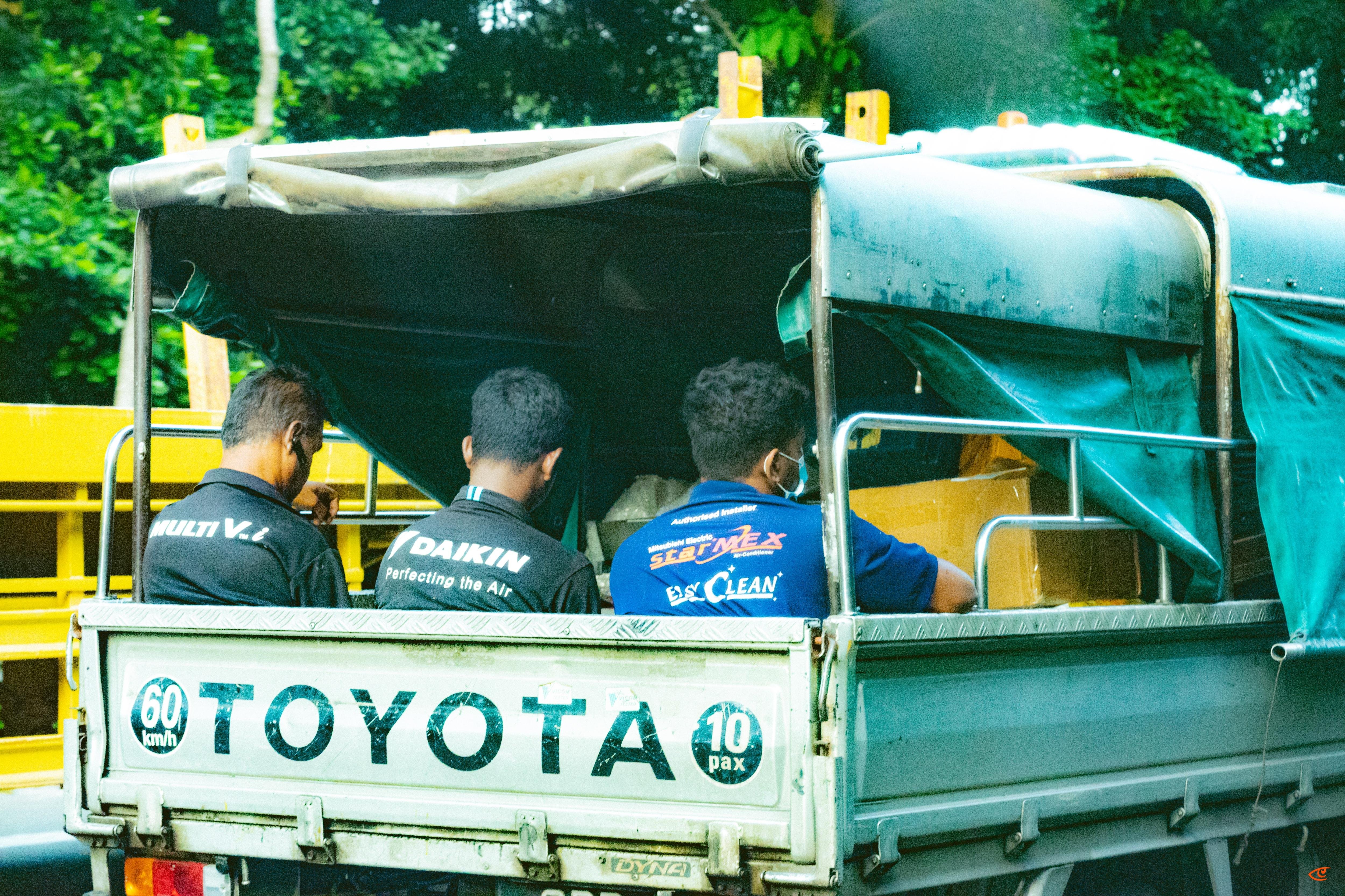 Three men seated with their backs to the camera in the open rear of a Toyota pickup truck, partially covered by a tarp canopy; “TOYOTA” printed on the tailgate with “60 km/h” and “10 pax” stickers, yellow railings and green trees in the background.