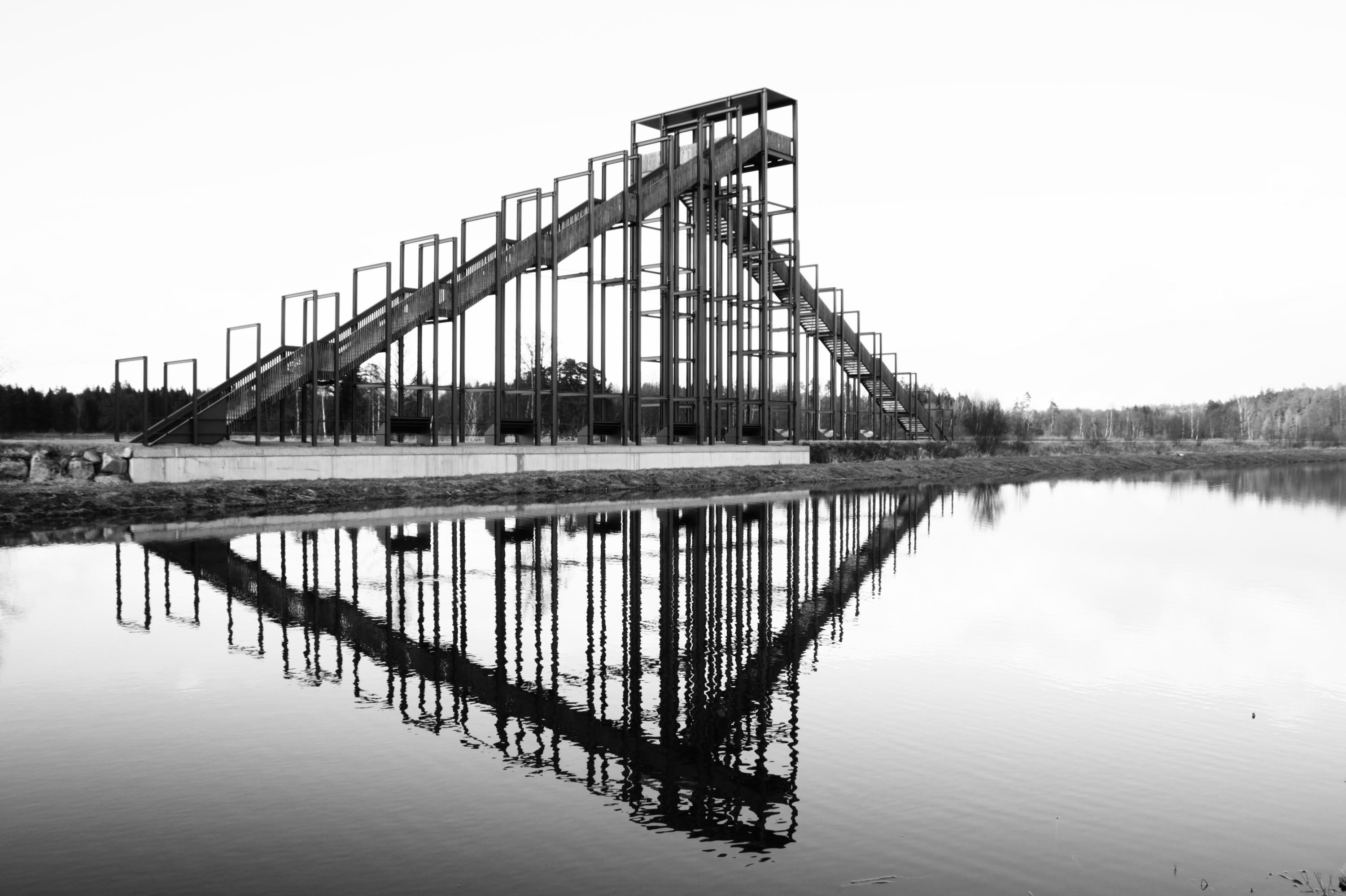 Black-and-white photo of a large metal staircase structure on a concrete base beside a calm lake, with the framework and steps reflected in the water; trees and low hills in the distant background.