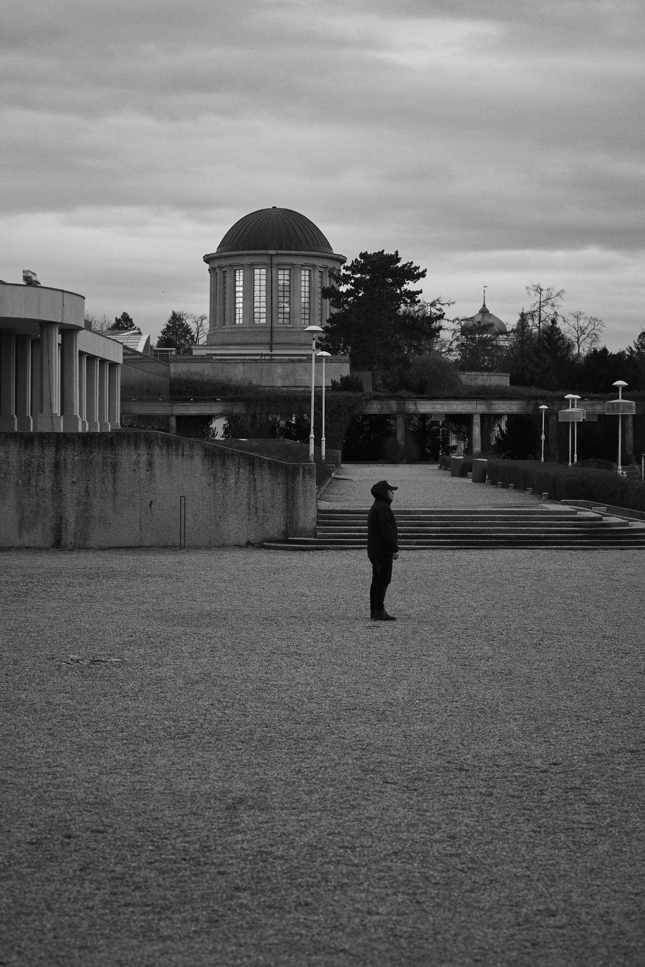 The image is a black and white photograph depicting a solitary person standing in an open area. In the background, there's a building with a dome and some trees. The setting appears to be spacious and architectural, possibly a park or a public area with pathways and structural features like walls and stairs.