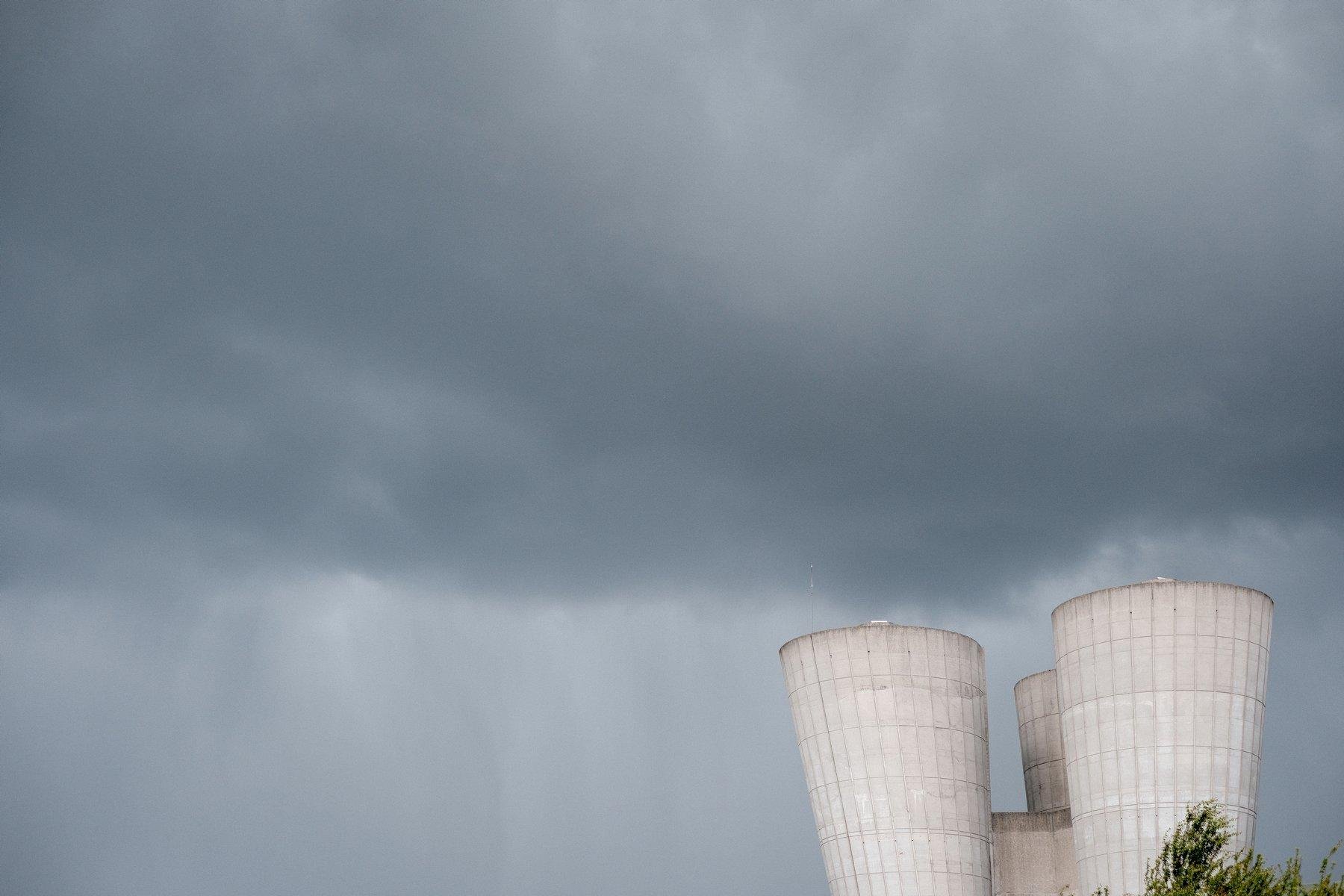 Photo horizontale. Majoritairement des nuages gris, foncés sur les 2/3 haut de l’image, puis une démarcation pour un gris plus clair. On devine un rideau de pluie. En bas à droite, une structure en béton presque blanche par éclairage (normalement grise) et un bout de feuillage d’arbre