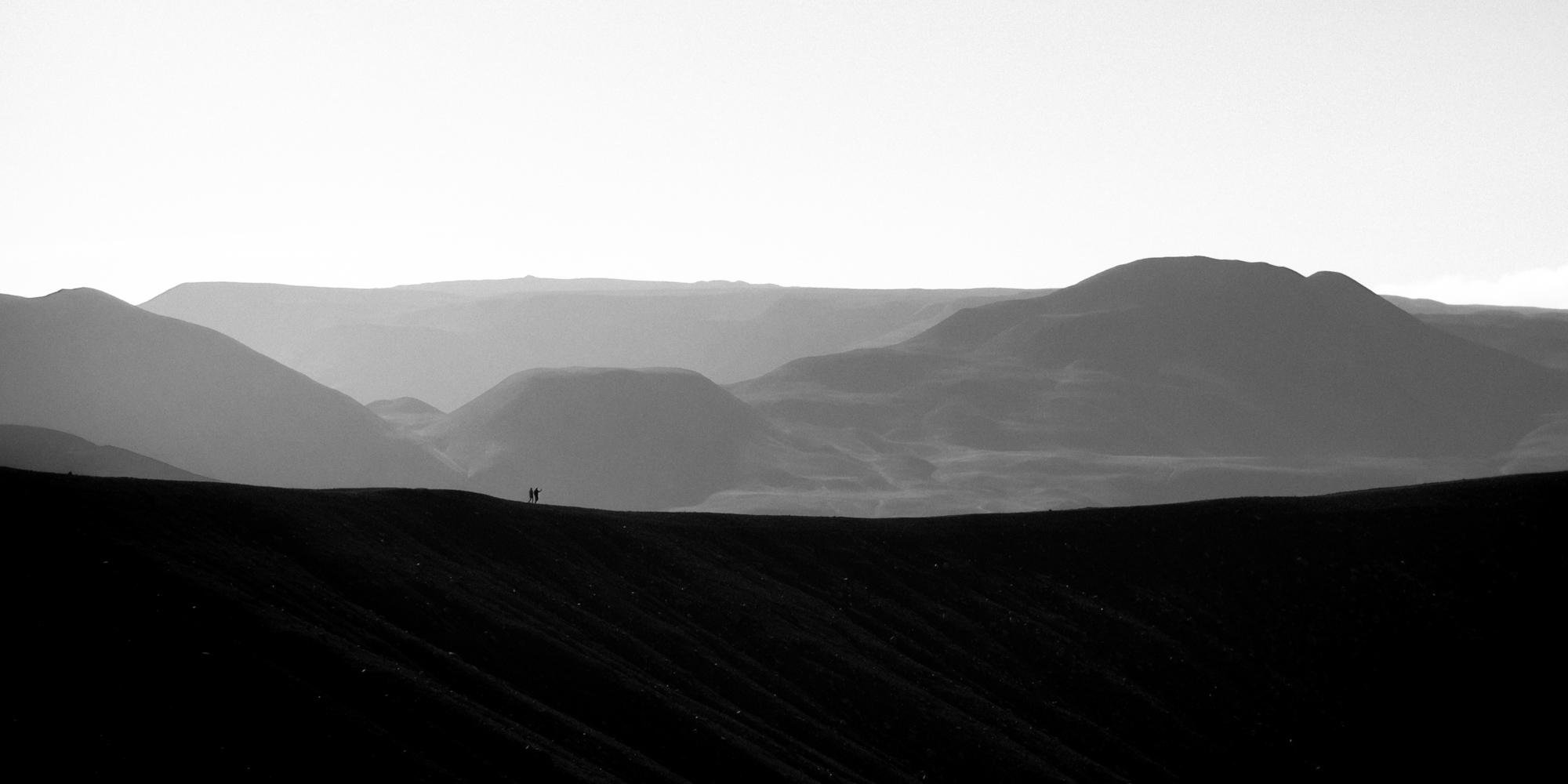 This is a black and white photograph showing a silhouette landscape of rolling hills in the background, with a dark ridge in the foreground. There appears to be a silhouette of two small figures on the ridge, suggesting a sense of scale and distance within the scene. The image has a high contrast between the dark foreground and the lighter, misty background.