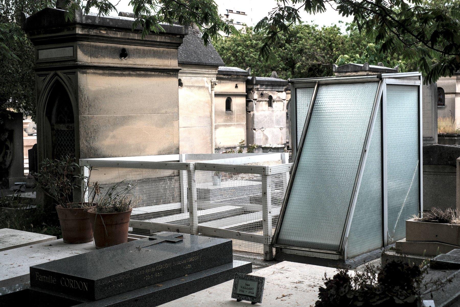 Photo horizontale. Au milieu des tombes, une sorte de petite cabine en verre à côté d’une parcelle délimité par un petit enclos blanc grillagé